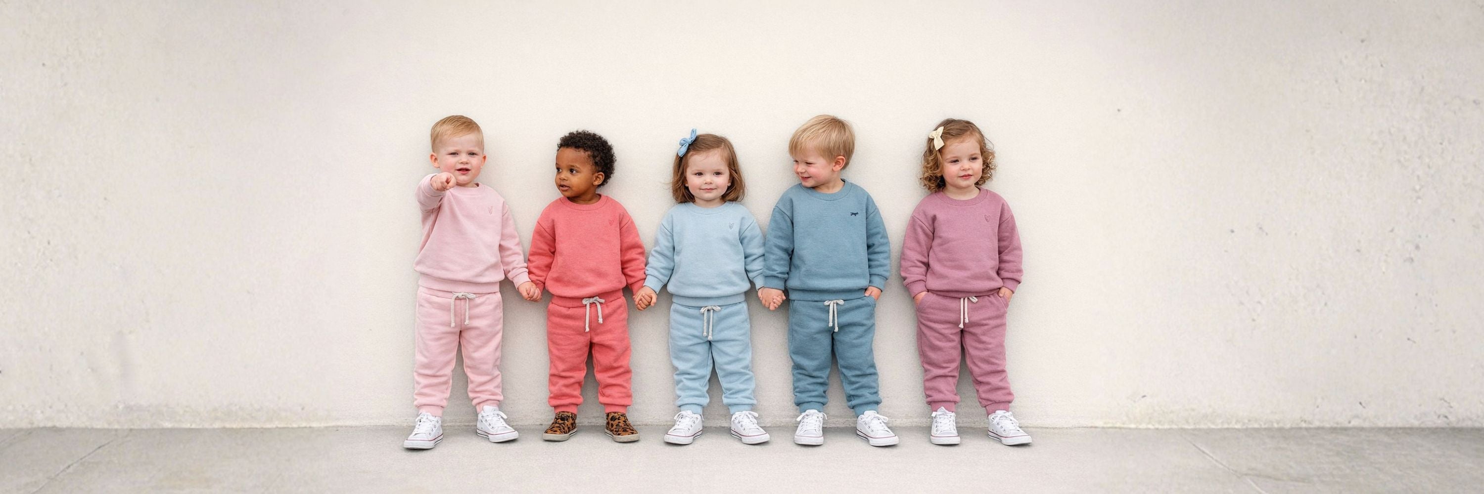 Five toddlers standing side by side against a neutral background, holding hands and wearing matching tracksuits in soft pastel colors including pink, coral, light blue, and mauve, paired with casual sneakers.