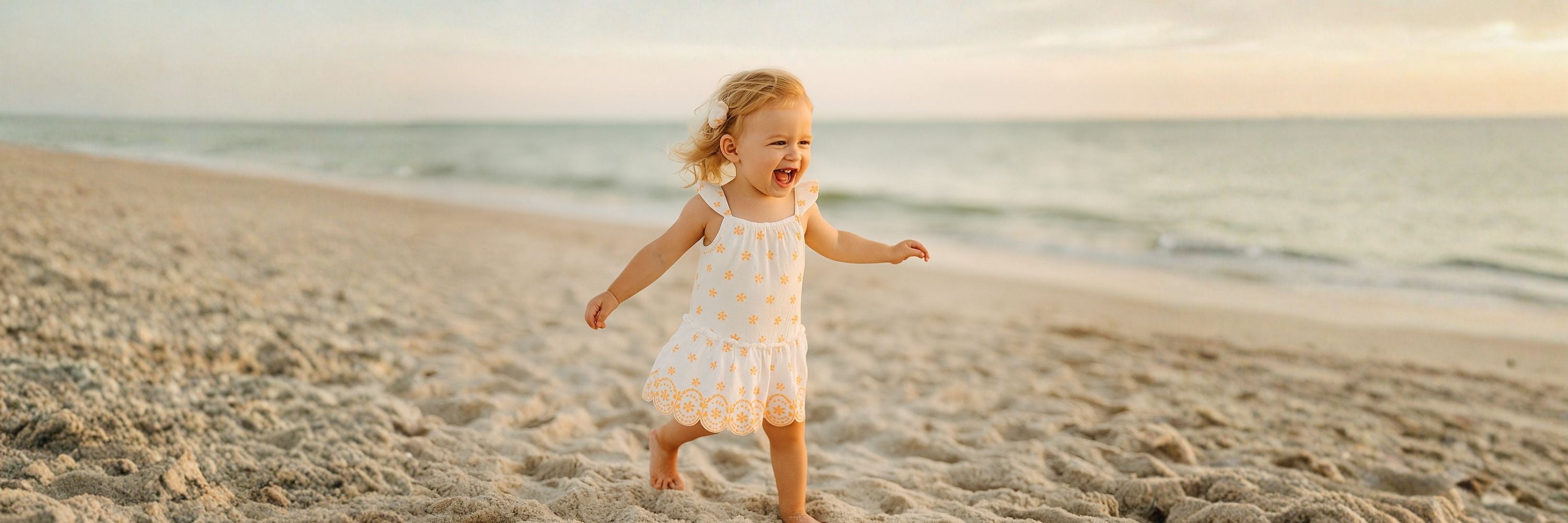 A smiling toddler running on the beach, wearing a light summer dress, with the sea and sandy shore in the background at sunset.