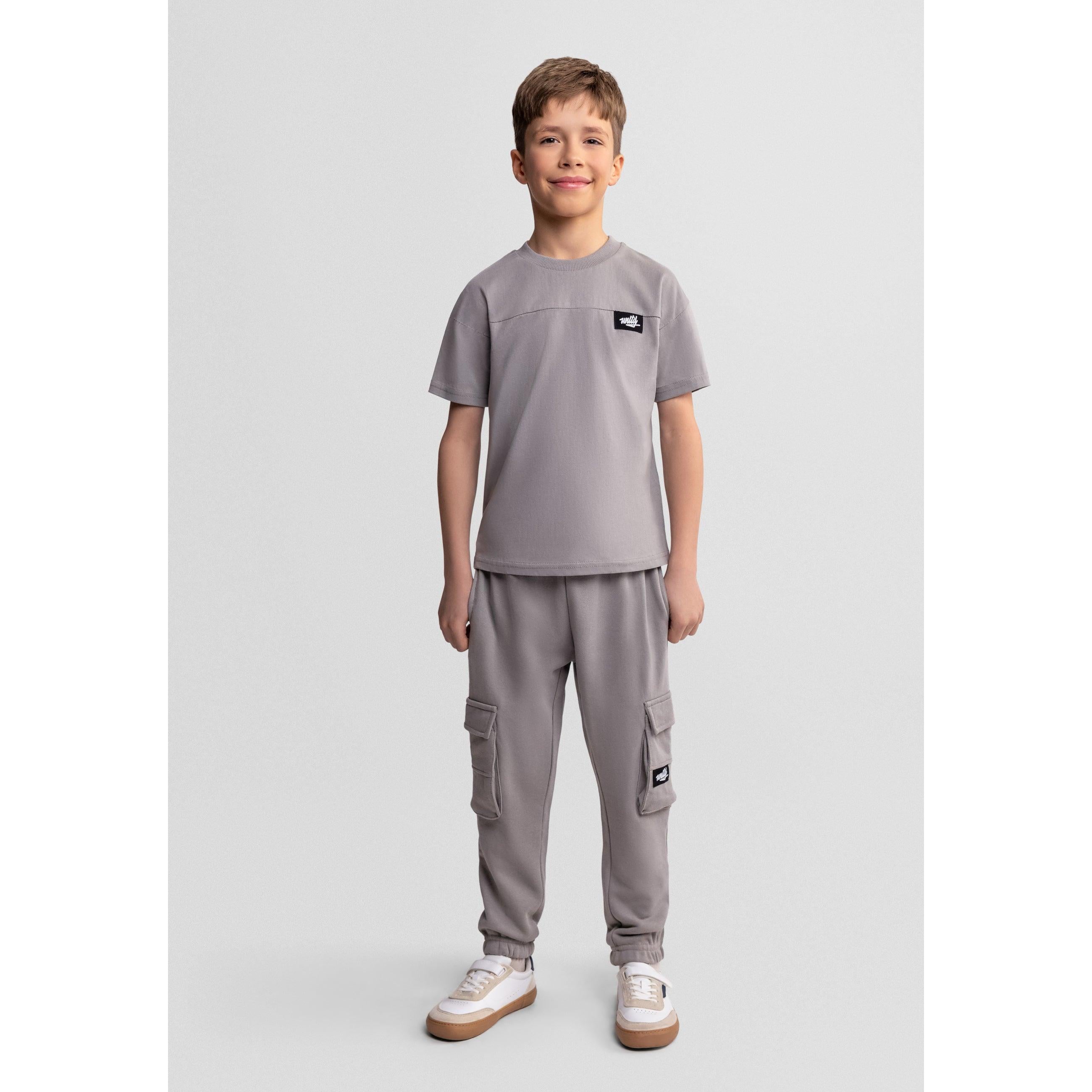 Young boy wearing grey jogging set with t-shirt and cargo joggers, standing against light grey background in white sneakers