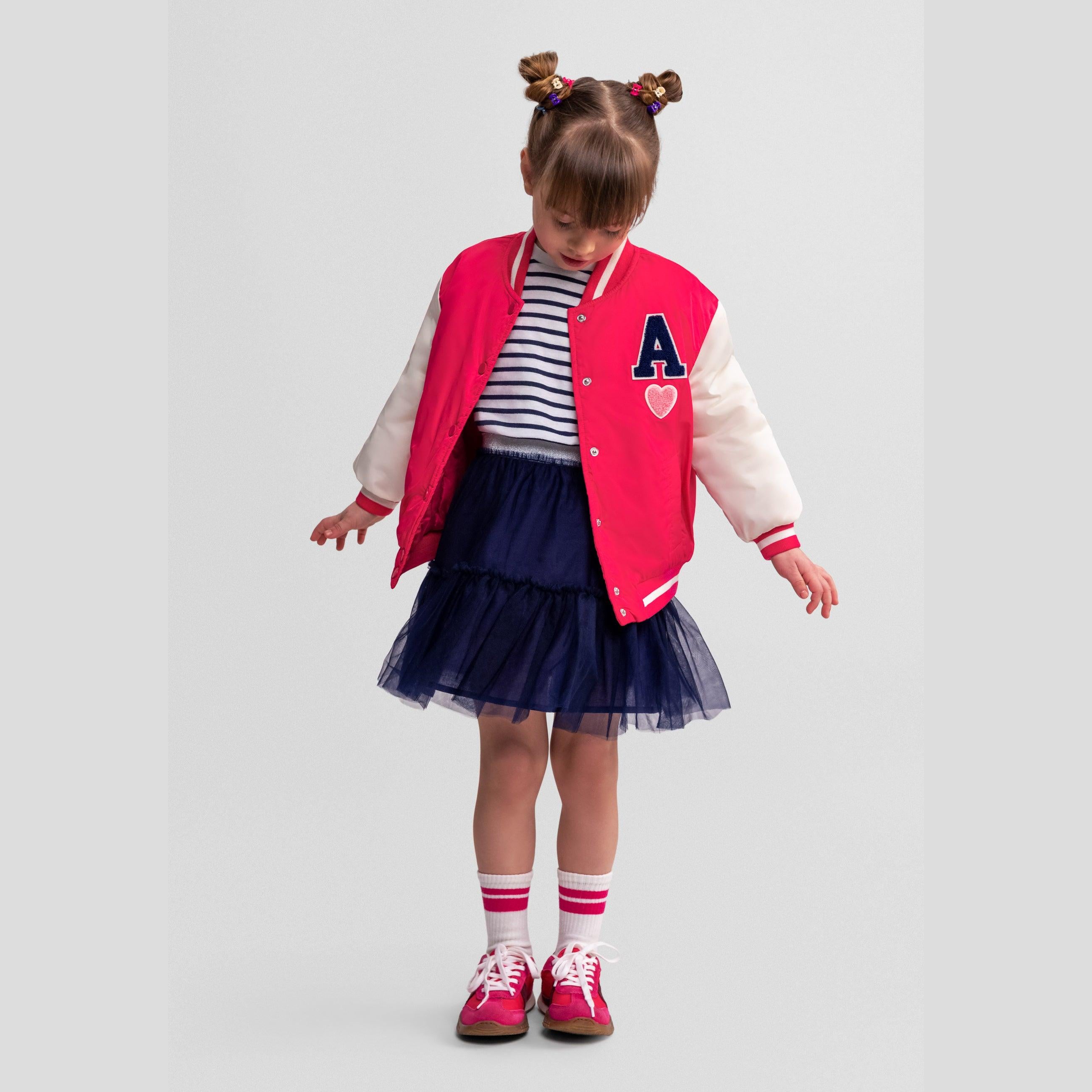 Young girl wearing pink varsity bomber jacket with navy tulle skirt and striped top, standing against white background