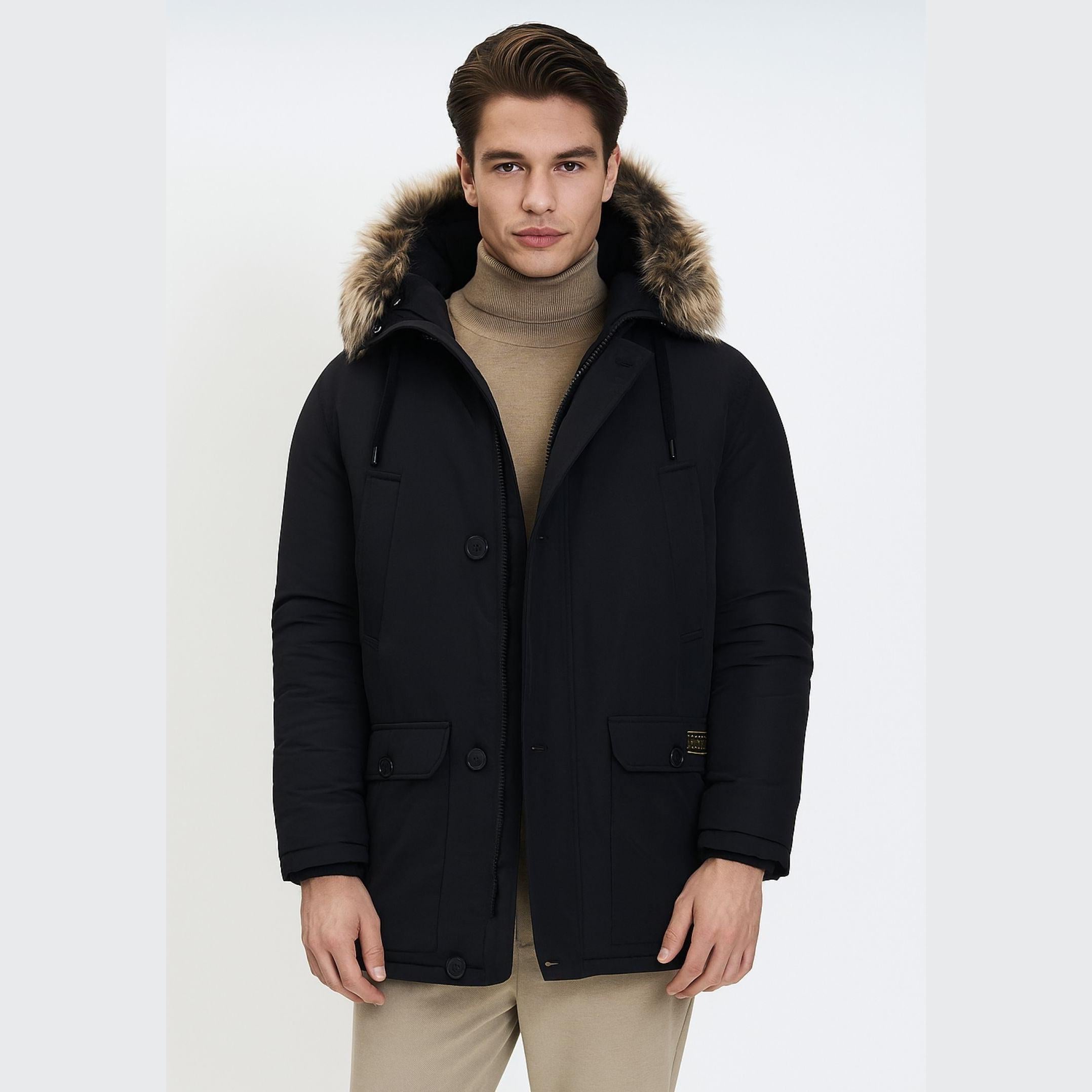 Young man wearing black padded parka jacket with faux fur hood trim over beige turtleneck sweater, standing against white background
