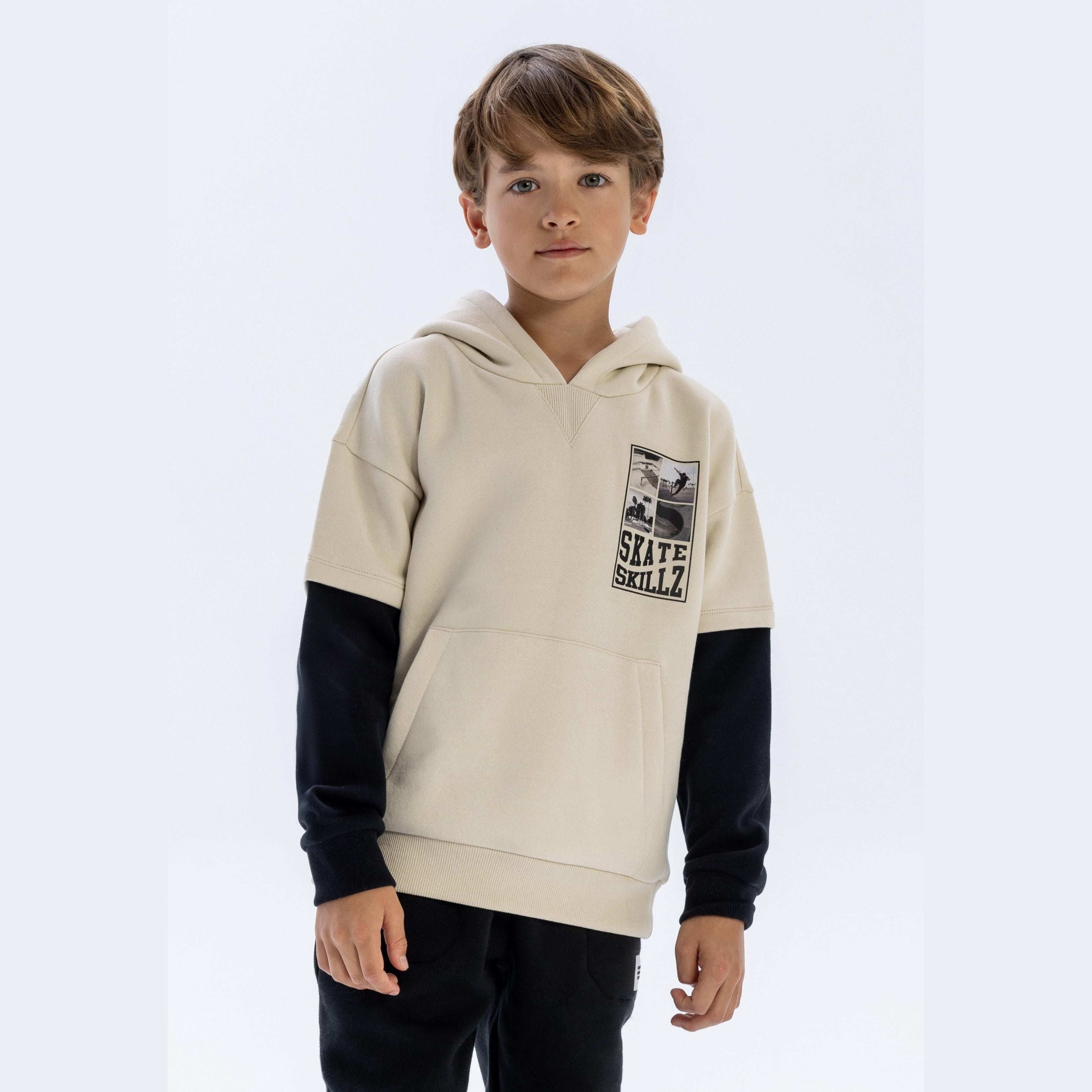 Young boy wearing stone hoodie with black sleeves and Skate Skillz print, standing against white background