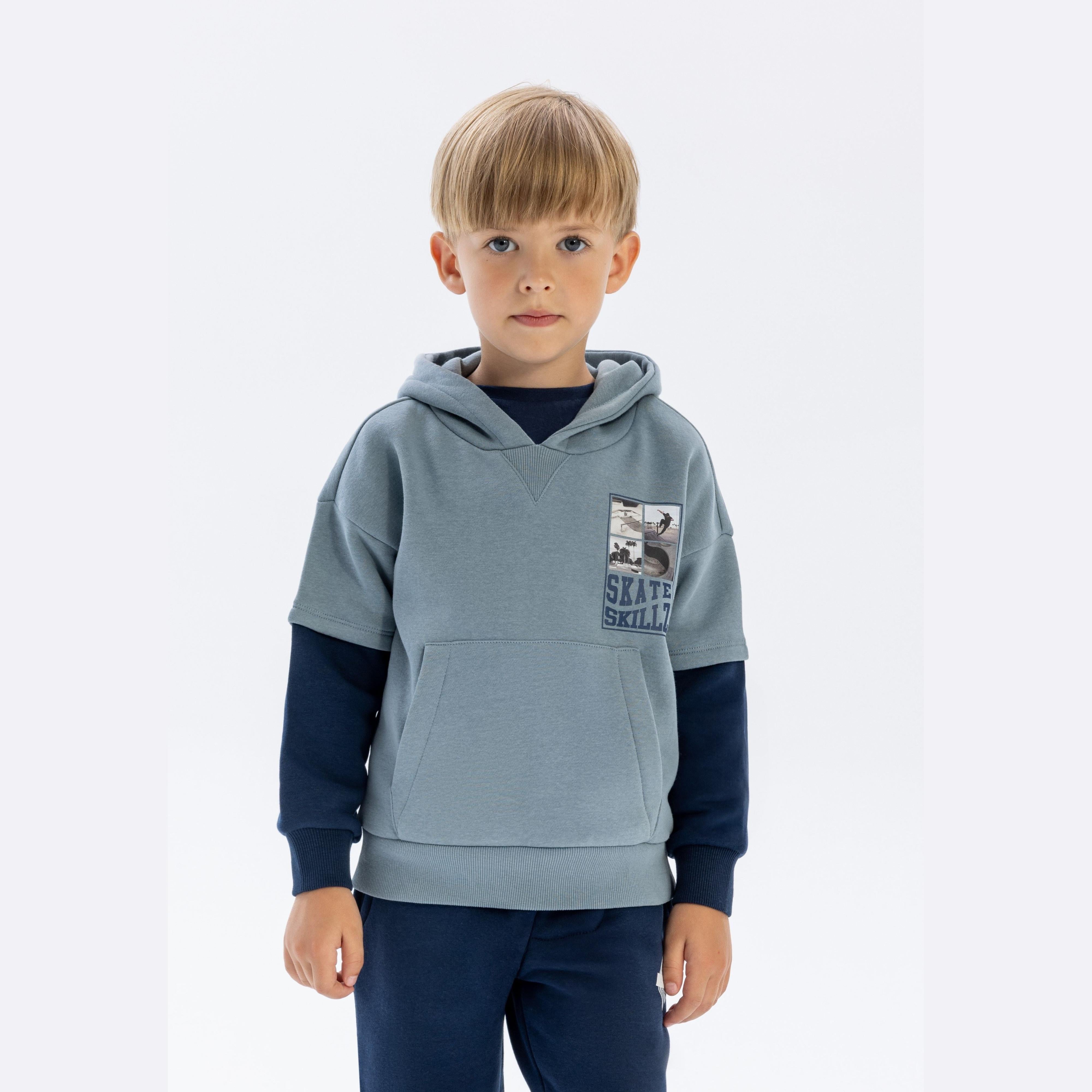 Young boy wearing blue hoodie with navy contrast sleeves and skateboarding graphics, standing against white background