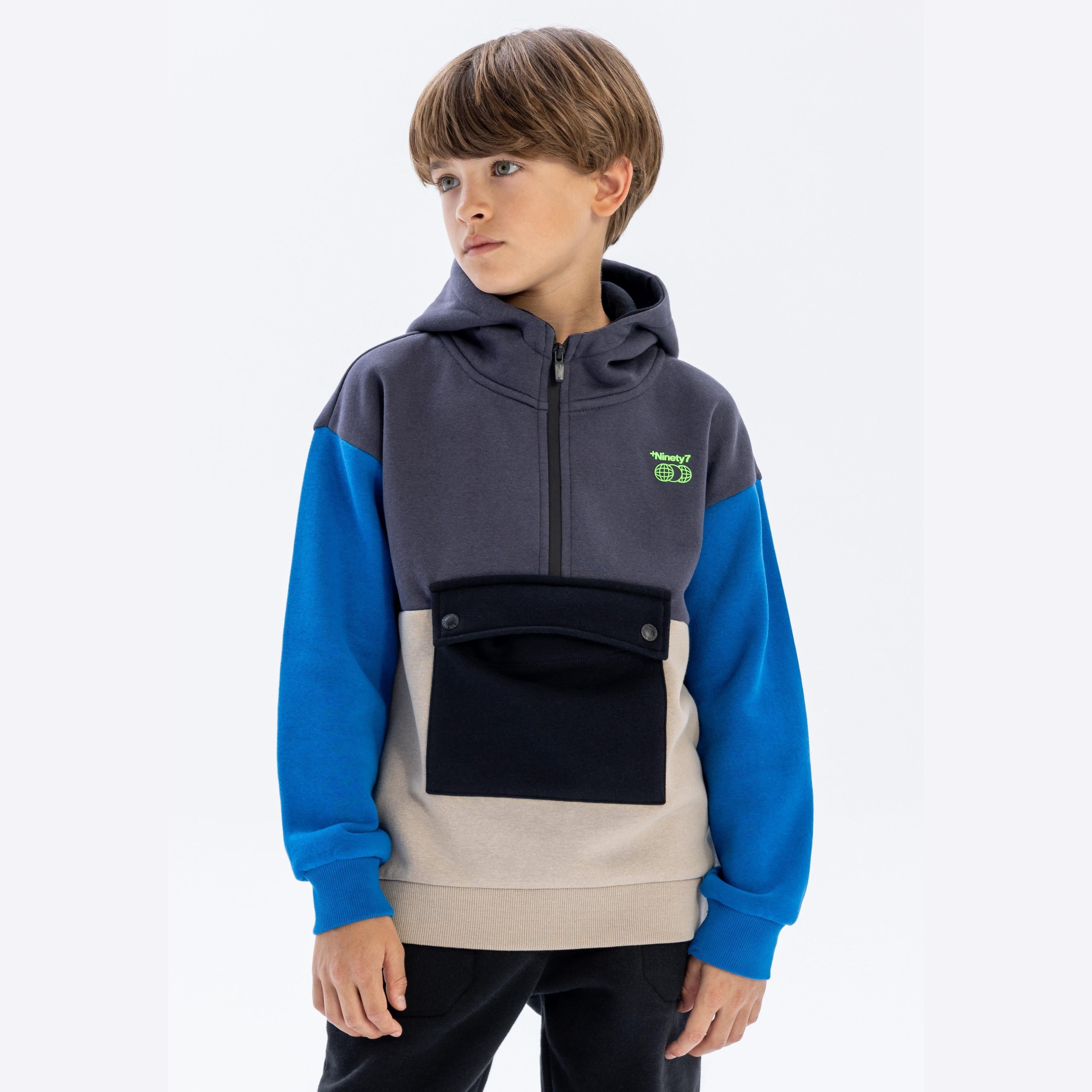 Young boy wearing grey, blue and cream colourblock hoodie with half zip, standing against white background