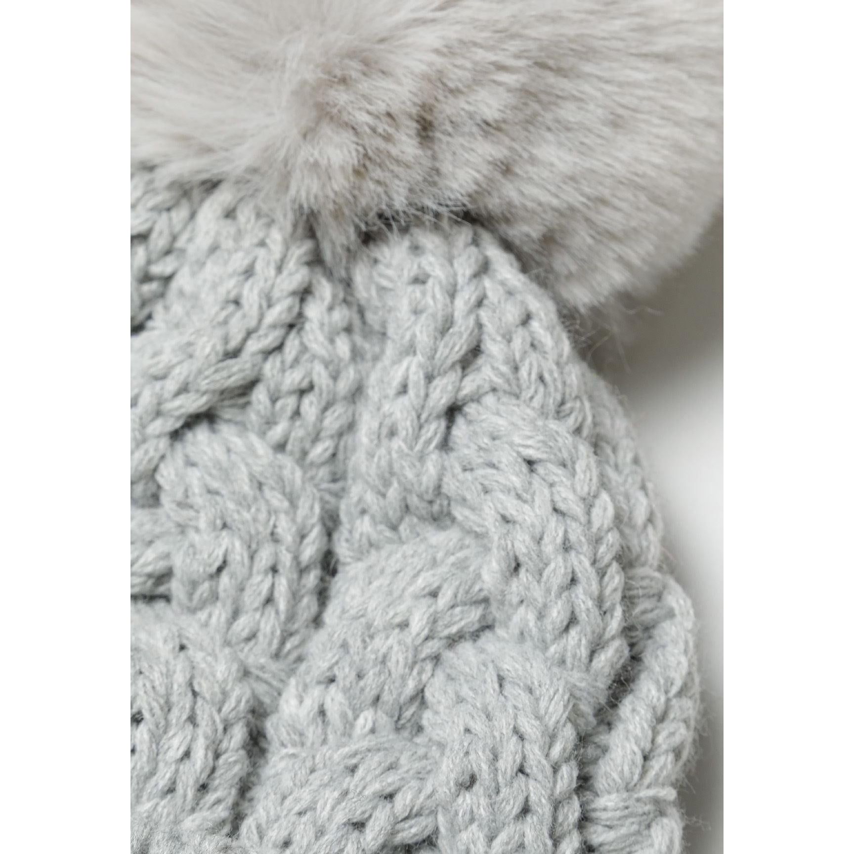 Close-up detail of grey cable knit pattern and fluffy pom pom attachment, showing soft knitted texture and craftsmanship quality