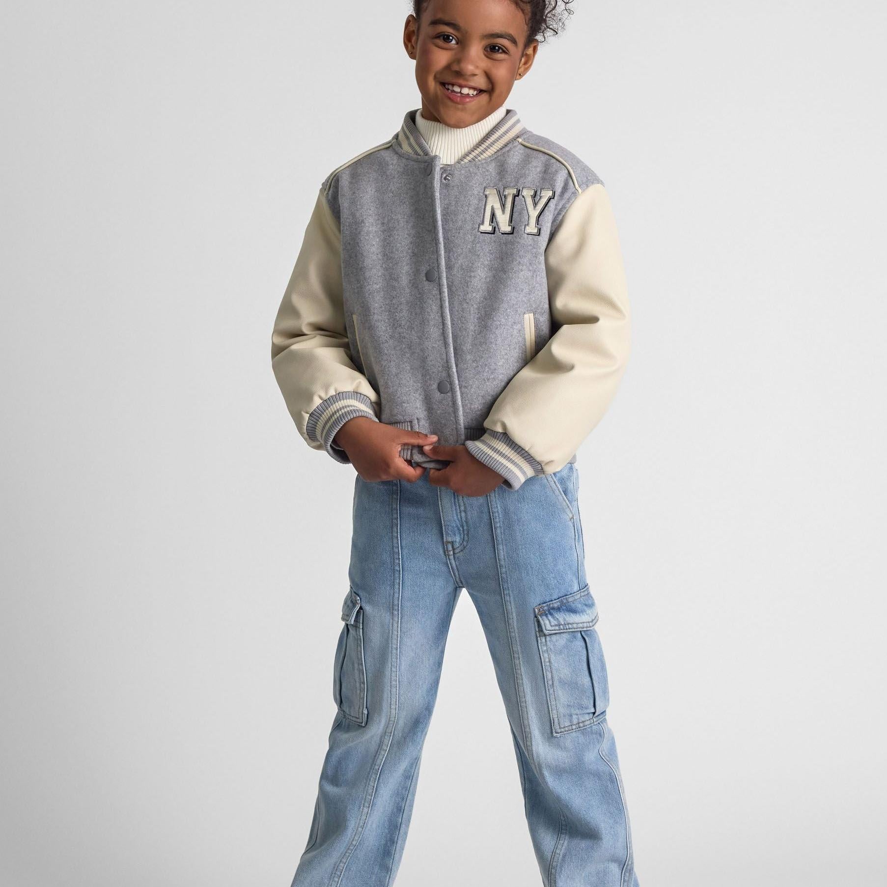 Young girl wearing grey baseball jacket with cream faux leather sleeves, paired with blue cargo jeans, standing against white background with hands in pockets