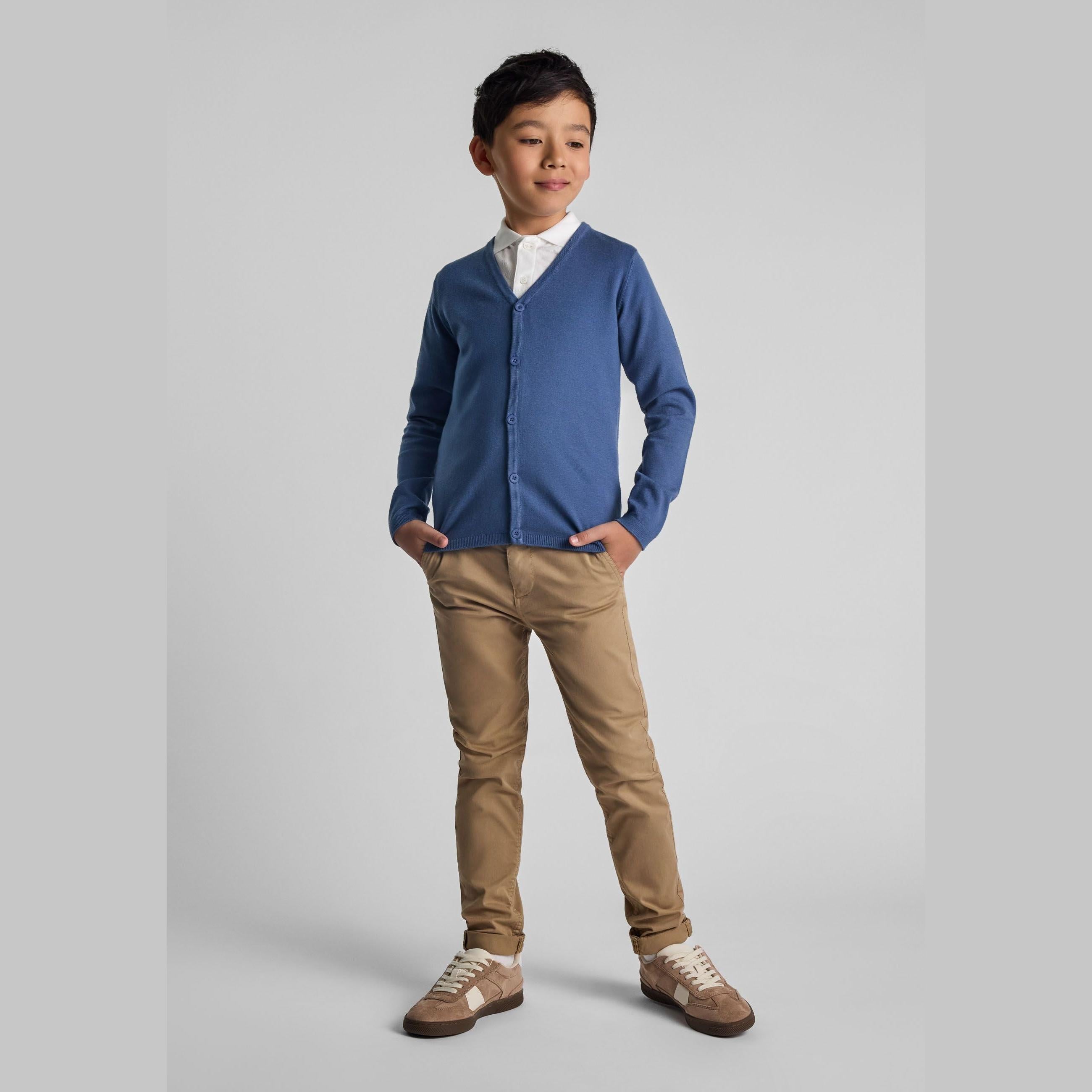 Young boy wearing blue button-down cardigan over white shirt with beige trousers, standing confidently against light grey studio background