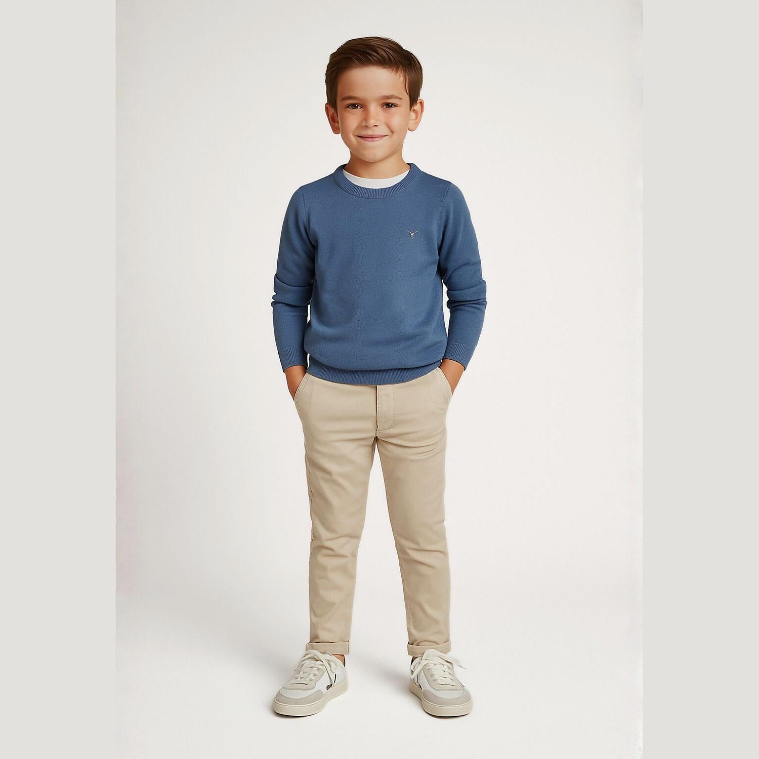 Boy wearing blue fine knit crew neck jumper with beige trousers and white sneakers, standing against light grey studio background