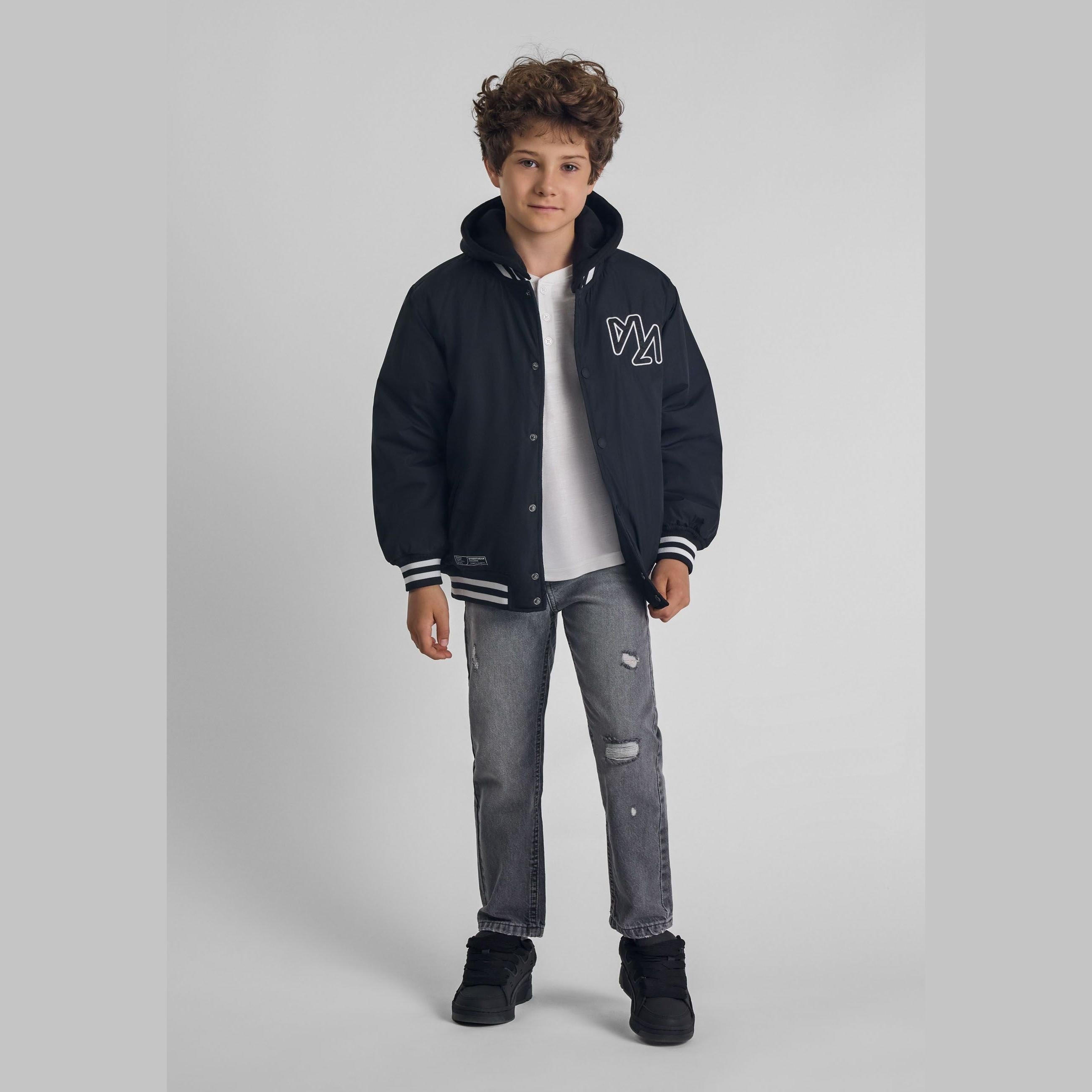 Young boy modeling grey distressed jeans with rip and repair details, wearing navy bomber jacket and white t-shirt, standing against neutral studio background