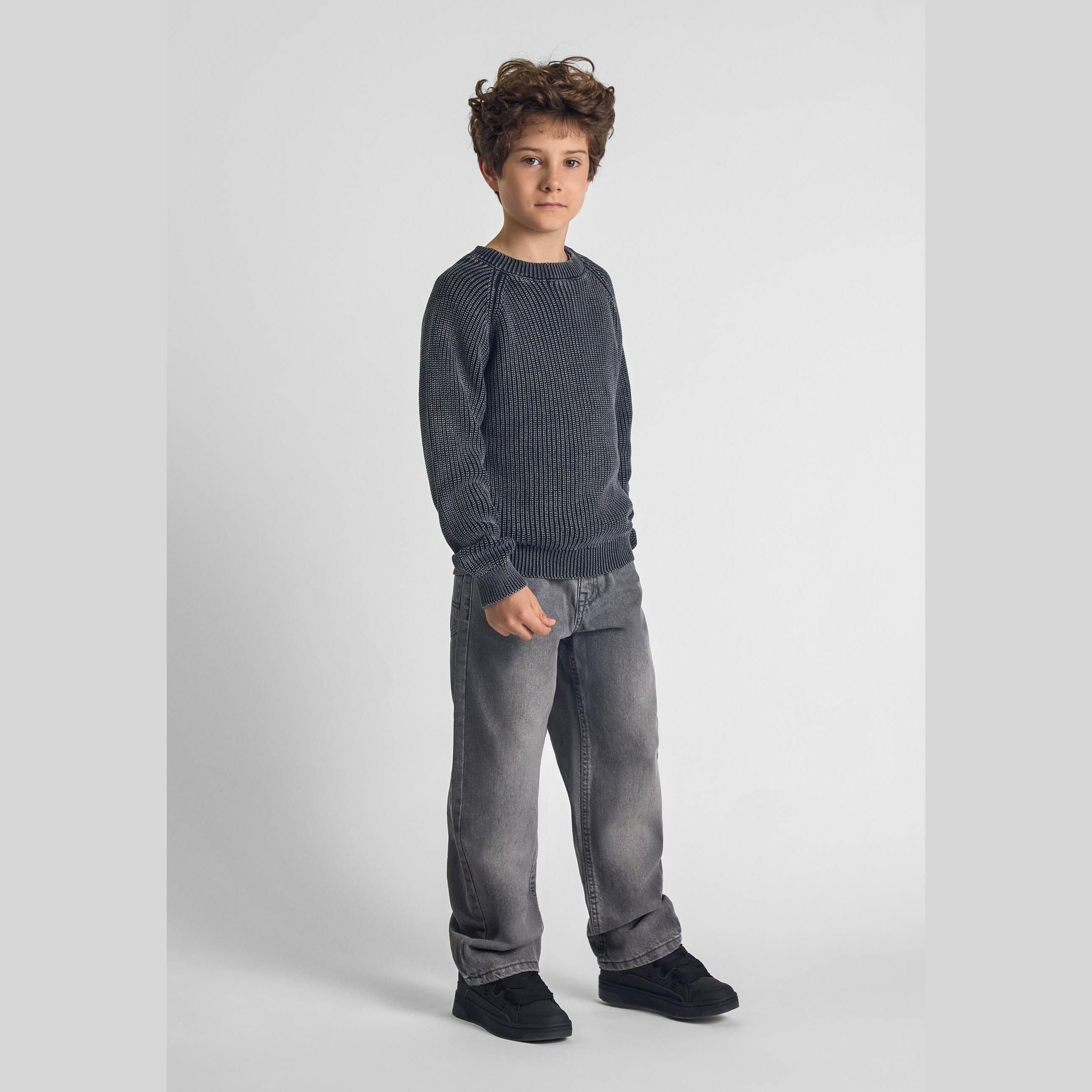 Boy wearing grey baggy jeans and dark knit sweater in studio setting with neutral background, full body view showing relaxed wide-leg denim fit