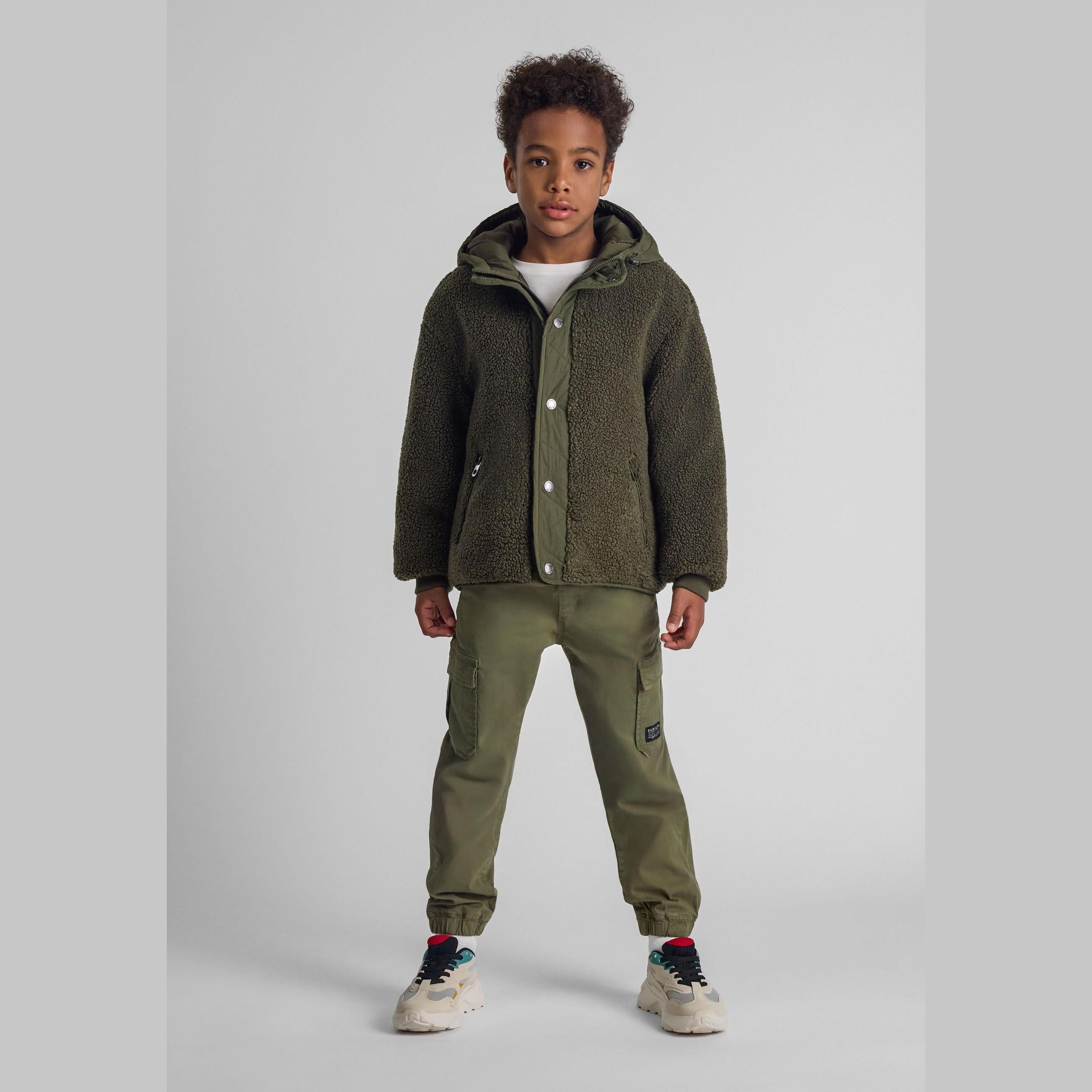 Young boy wearing khaki cargo trousers with elasticated waist and dark olive green teddy fleece jacket, standing against white background in studio setting