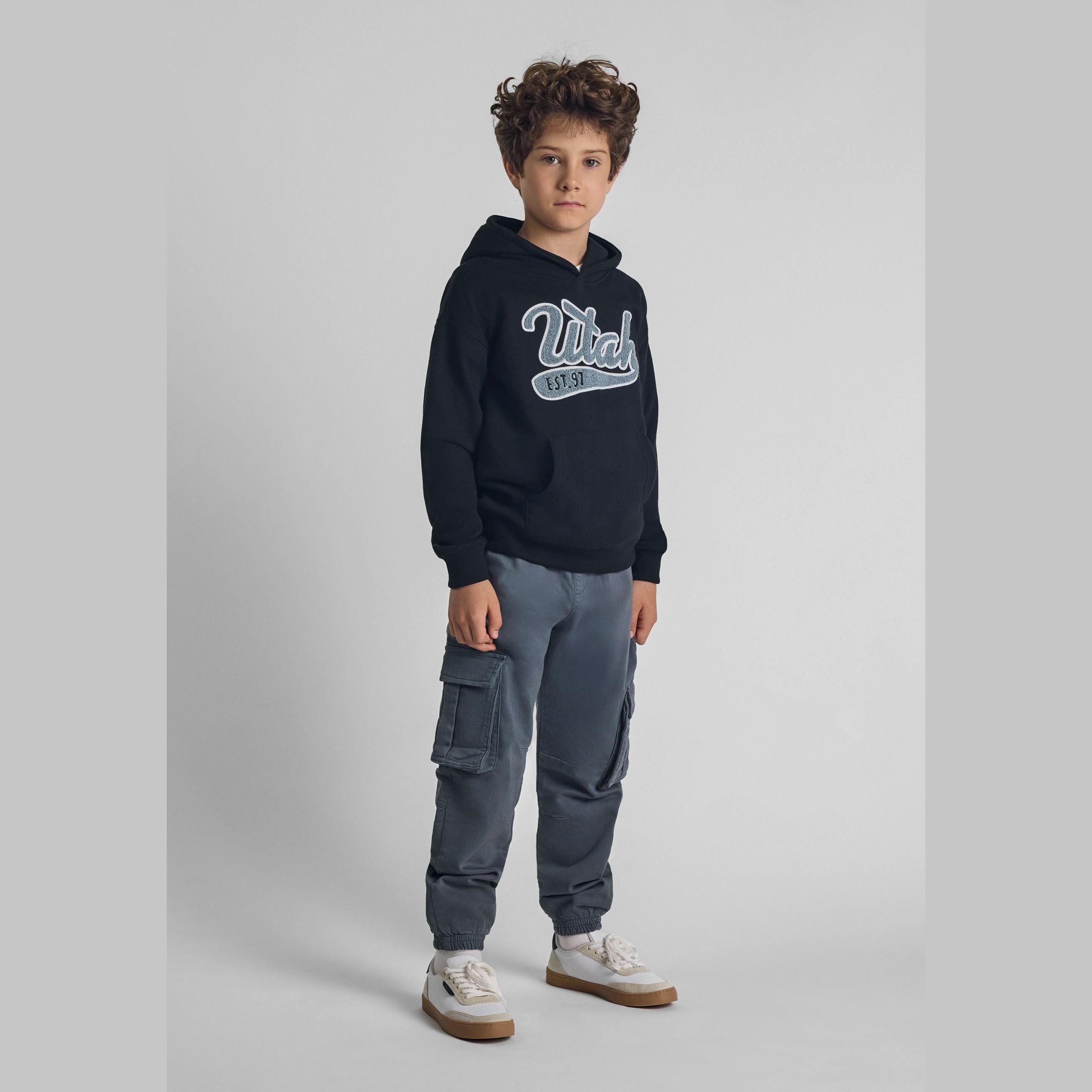 Boy wearing charcoal cargo trousers with navy hoodie against grey background showing full outfit styling