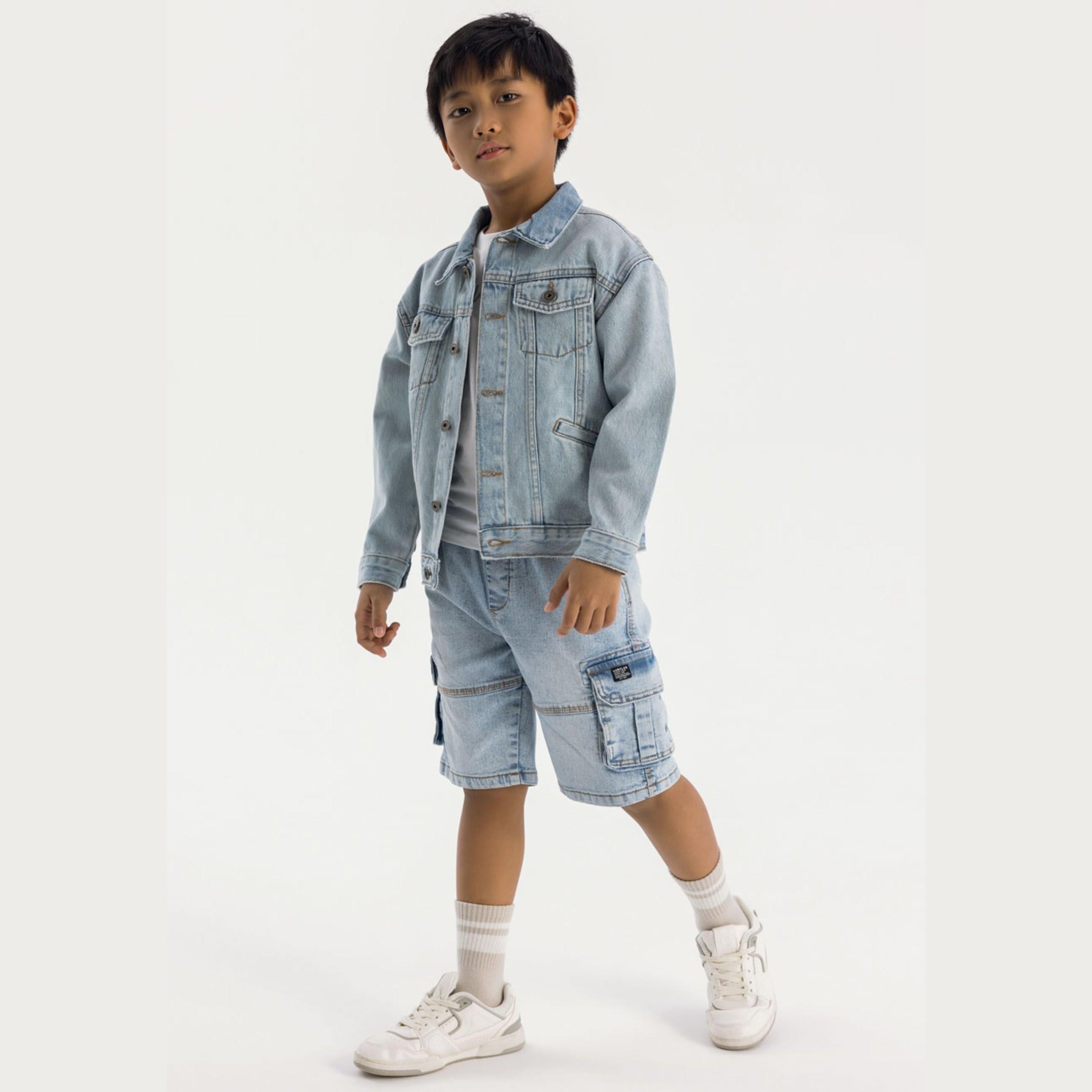 Young boy wearing light blue denim jacket and cargo shorts standing against white background, full body view showing casual spring styling