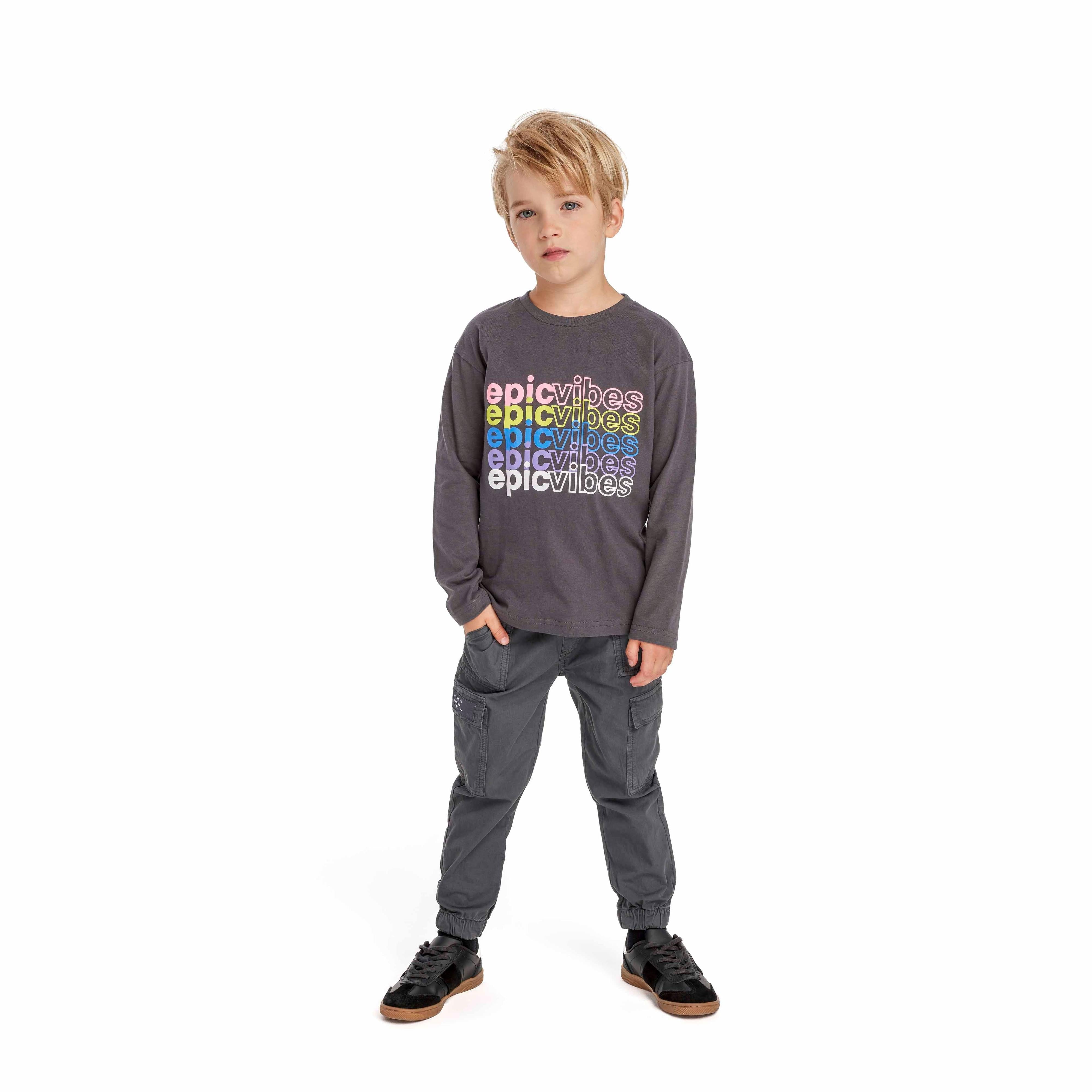 Young blonde boy wearing black combat trousers and grey long-sleeve top with colorful text, standing against white background with hands in pockets