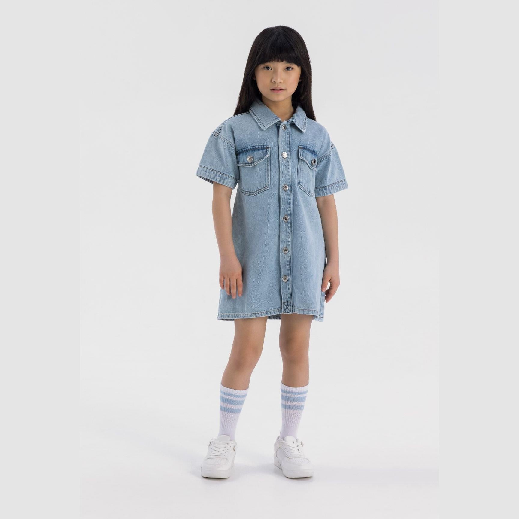 Young girl wearing light blue denim shirt dress with short sleeves, standing against white background with white sneakers and striped socks
