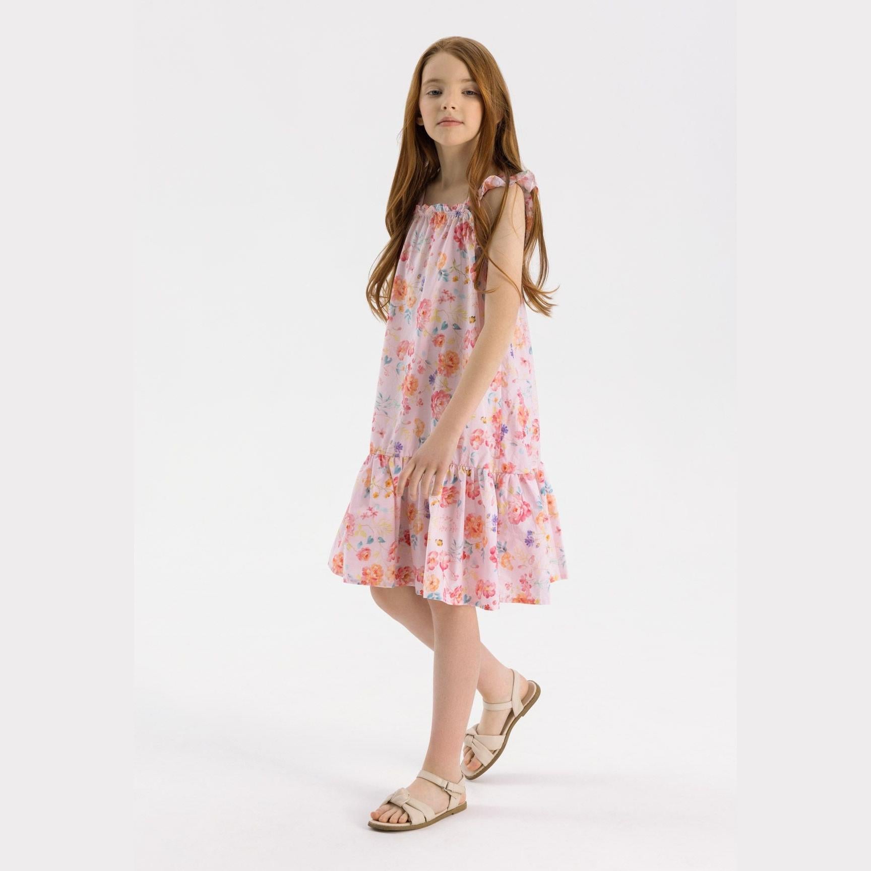 Young girl with long brown hair wearing pink floral cotton dress with ruffled straps and tiered skirt, standing against white background in beige sandals
