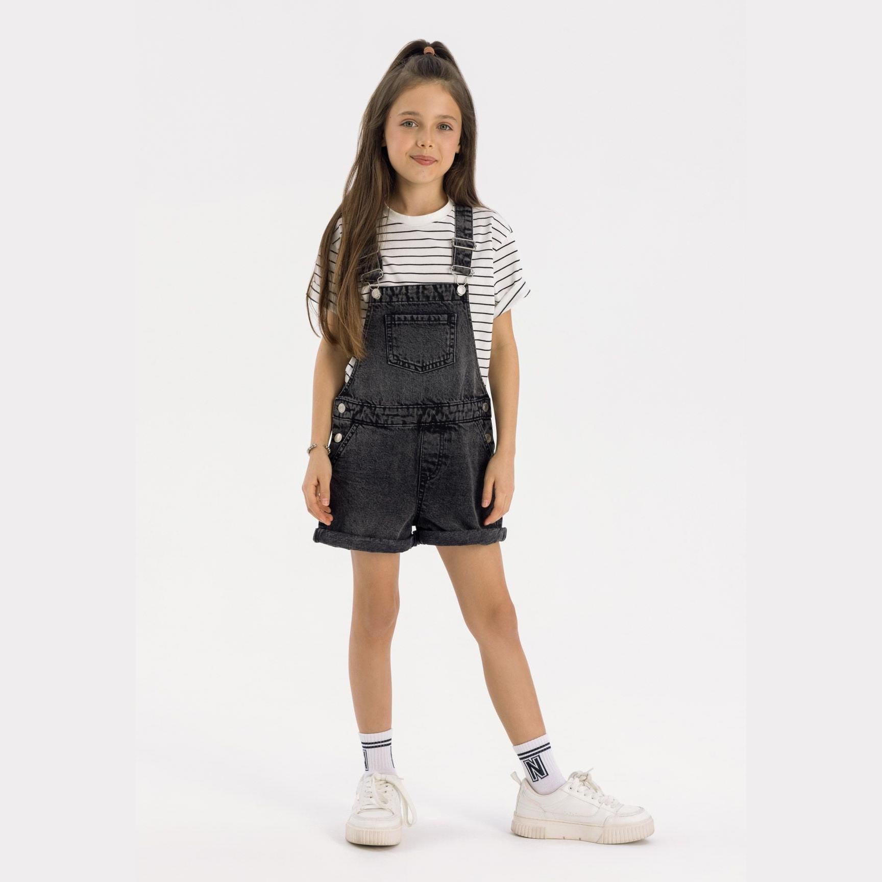 Young girl wearing black denim dungarees with striped top, white sneakers against light background