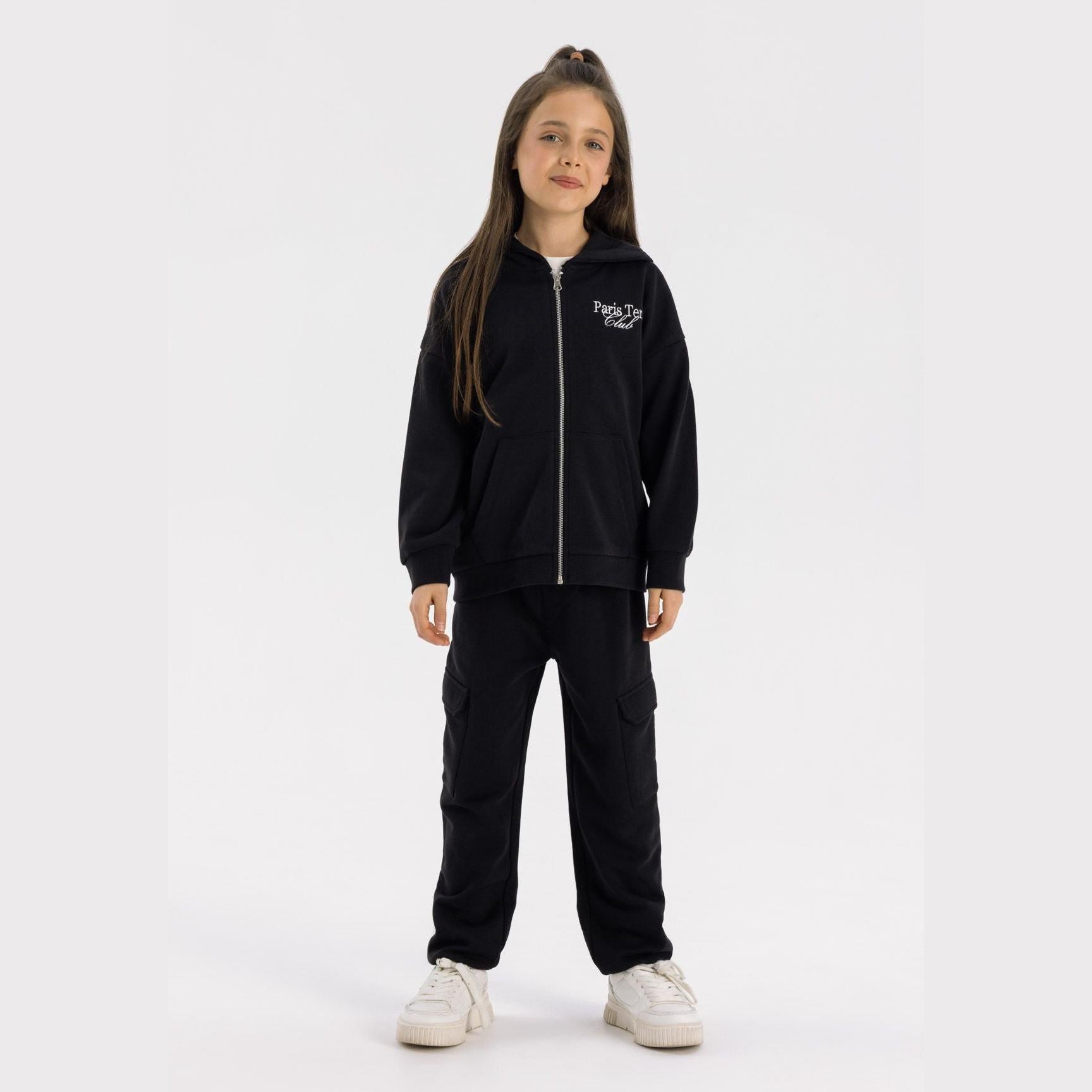 Young girl wearing black oversized zip hoodie with Paris Tennis Club embroidery and matching black joggers, standing against white background