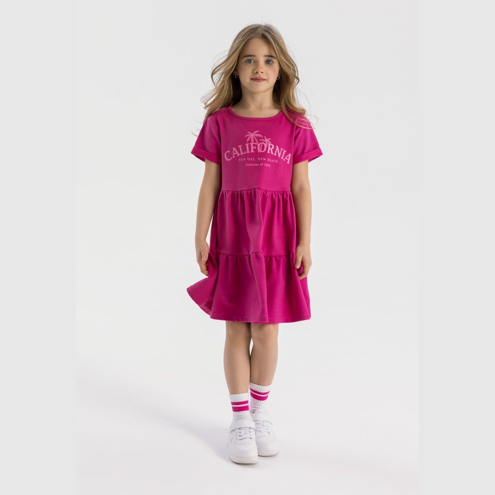 Young girl wearing pink cotton fleece tiered dress with California print, standing against white background, paired with white sneakers and striped socks