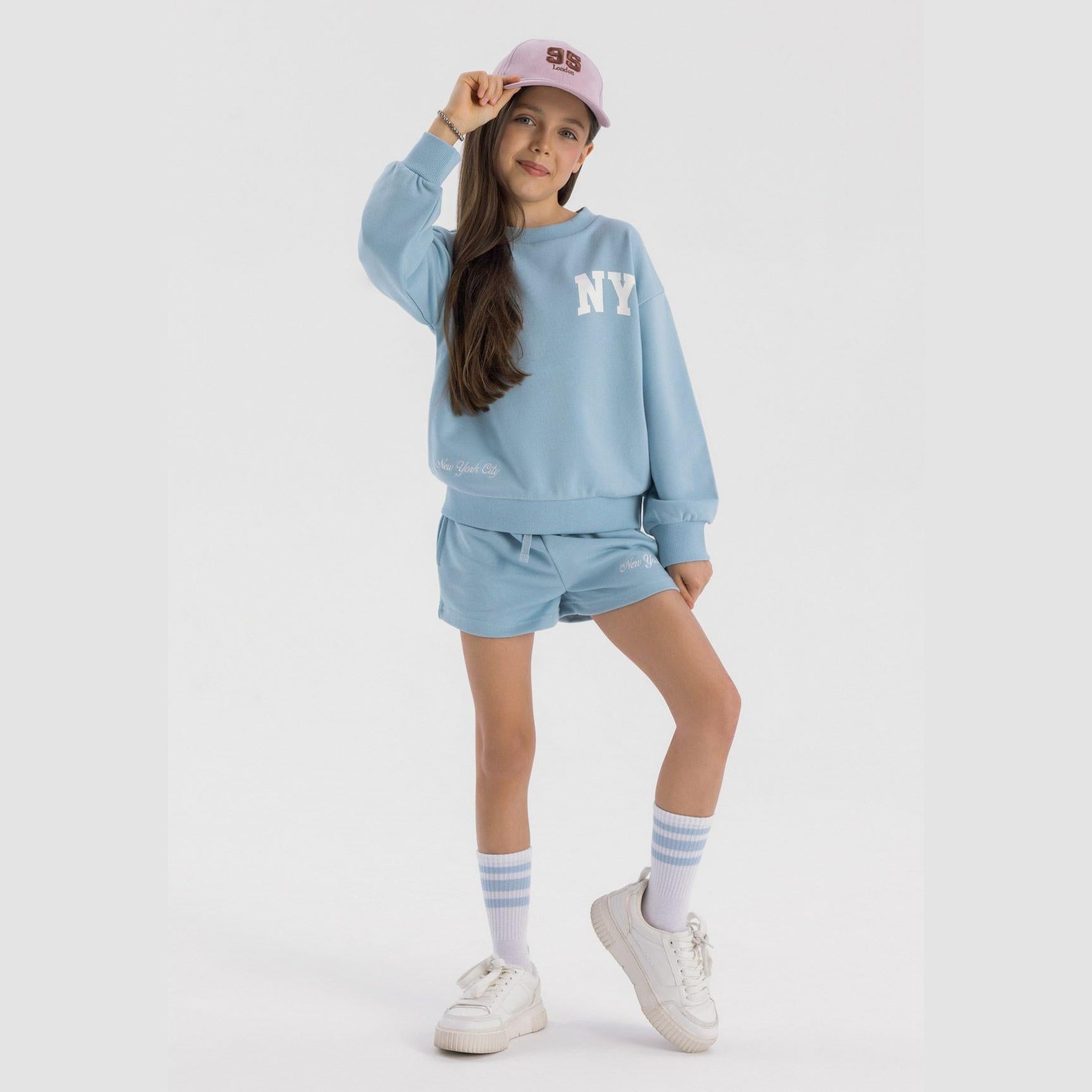 Young girl wearing blue crew neck sweatshirt and shorts set with NY print, pink baseball cap, white sneakers and striped socks against light grey background