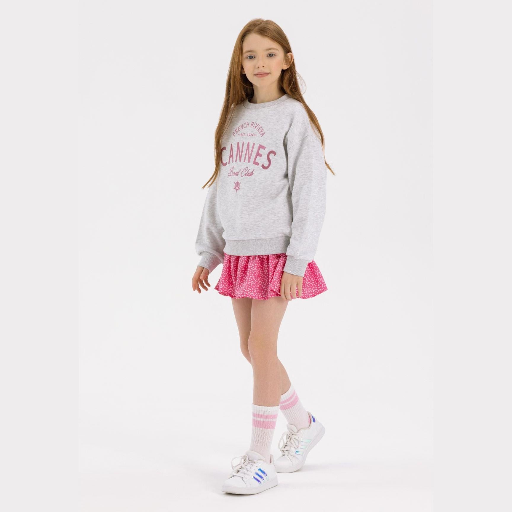 Young girl wearing grey CANNES sweatshirt and pink floral crinkle layered skirt, standing against white background with long brown hair and white sneakers