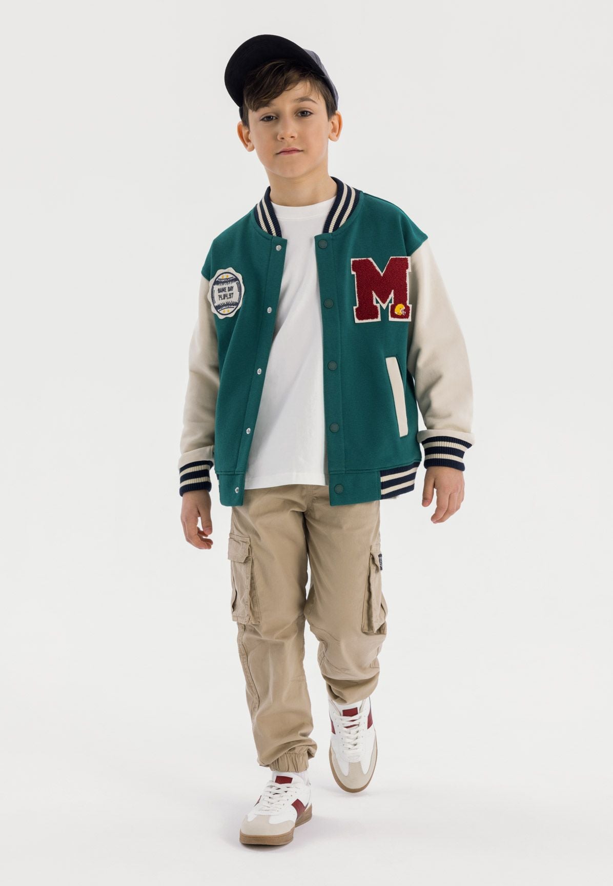 Young boy wearing green baseball jacket with cream sleeves and beige cargo pants, standing against white background