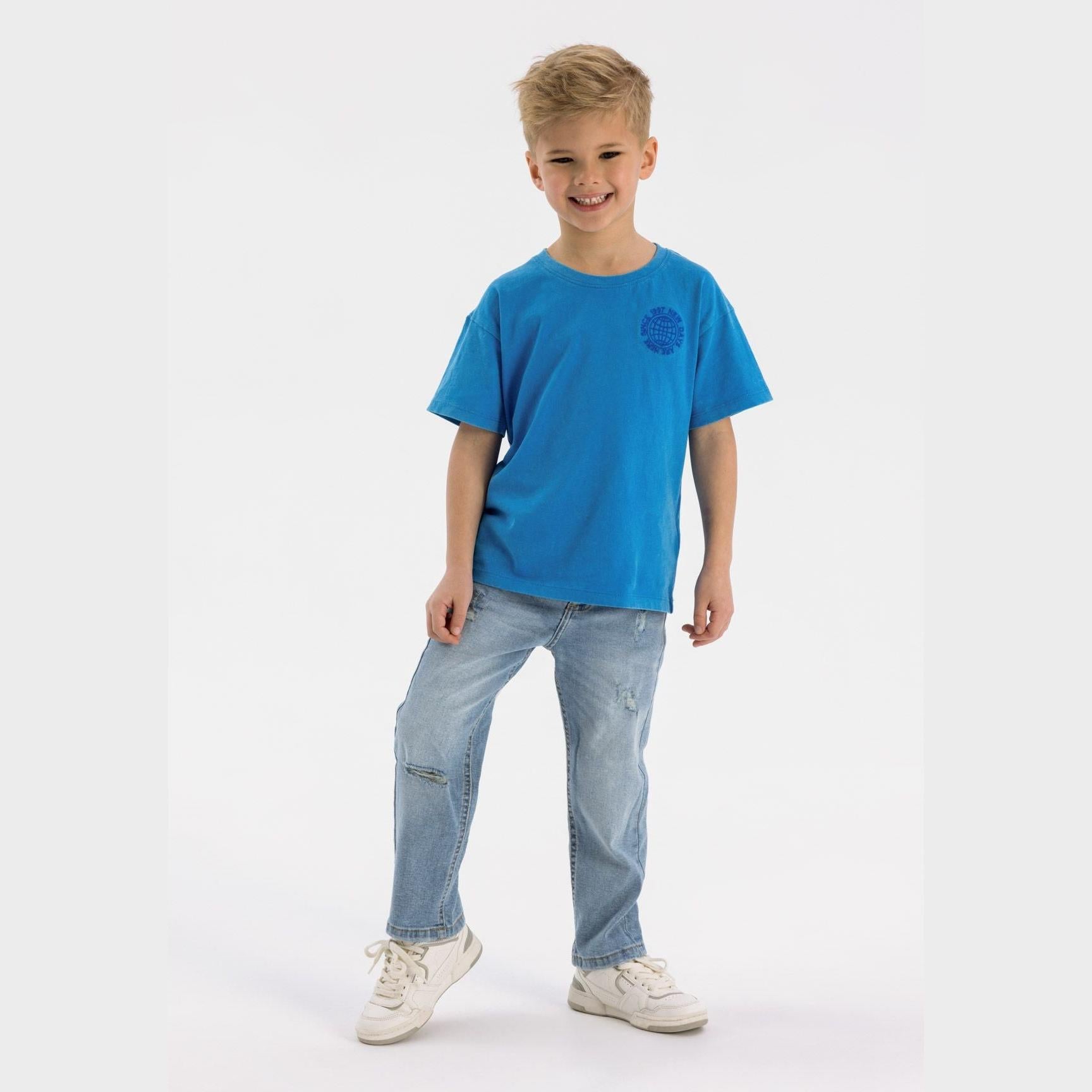 Young blonde boy wearing light blue ripped denim jeans with blue t-shirt, standing on white background with casual white sneakers