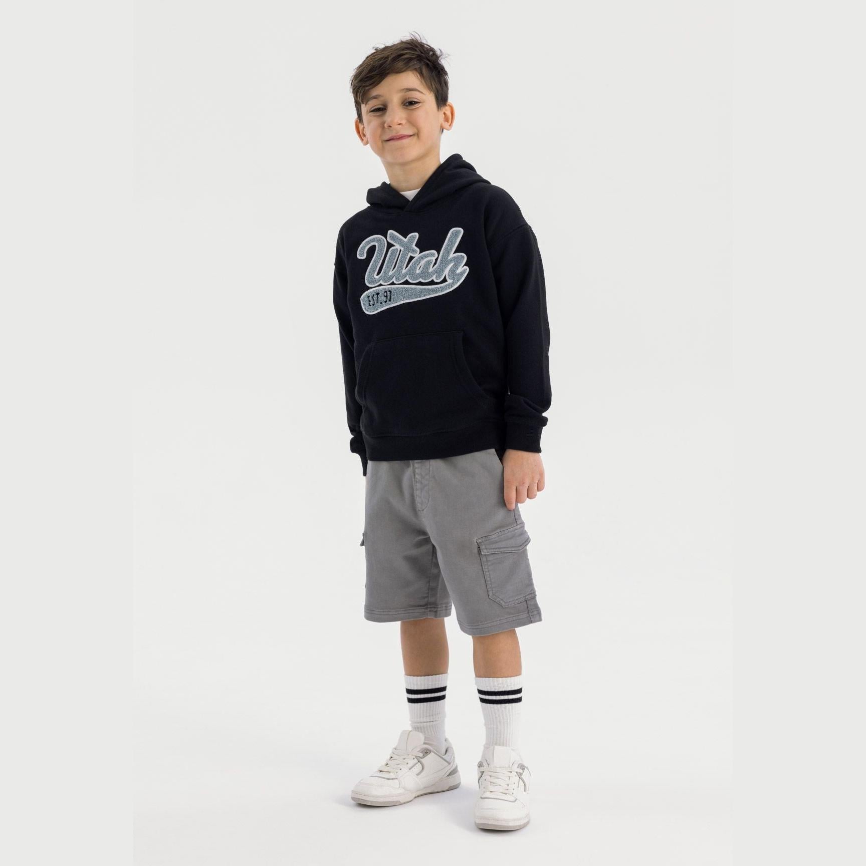 Young boy wearing black hoodie with Minoti logo and grey cargo shorts, standing against white background with white sneakers and striped socks