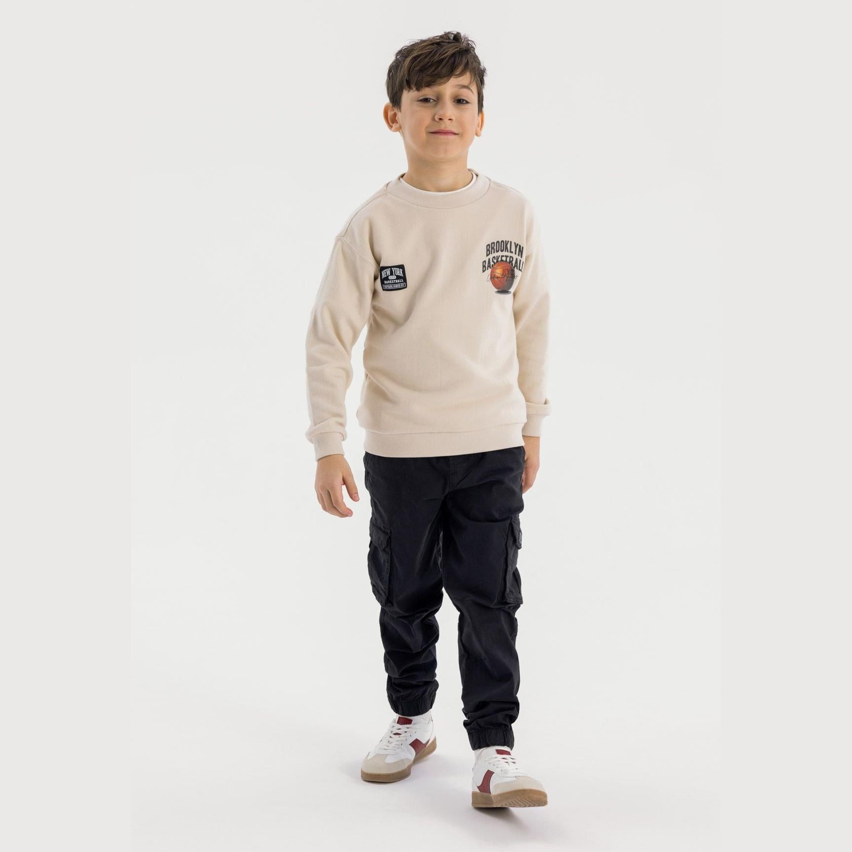Young boy wearing black cargo trousers with beige sweatshirt, standing on white background showing full outfit styling