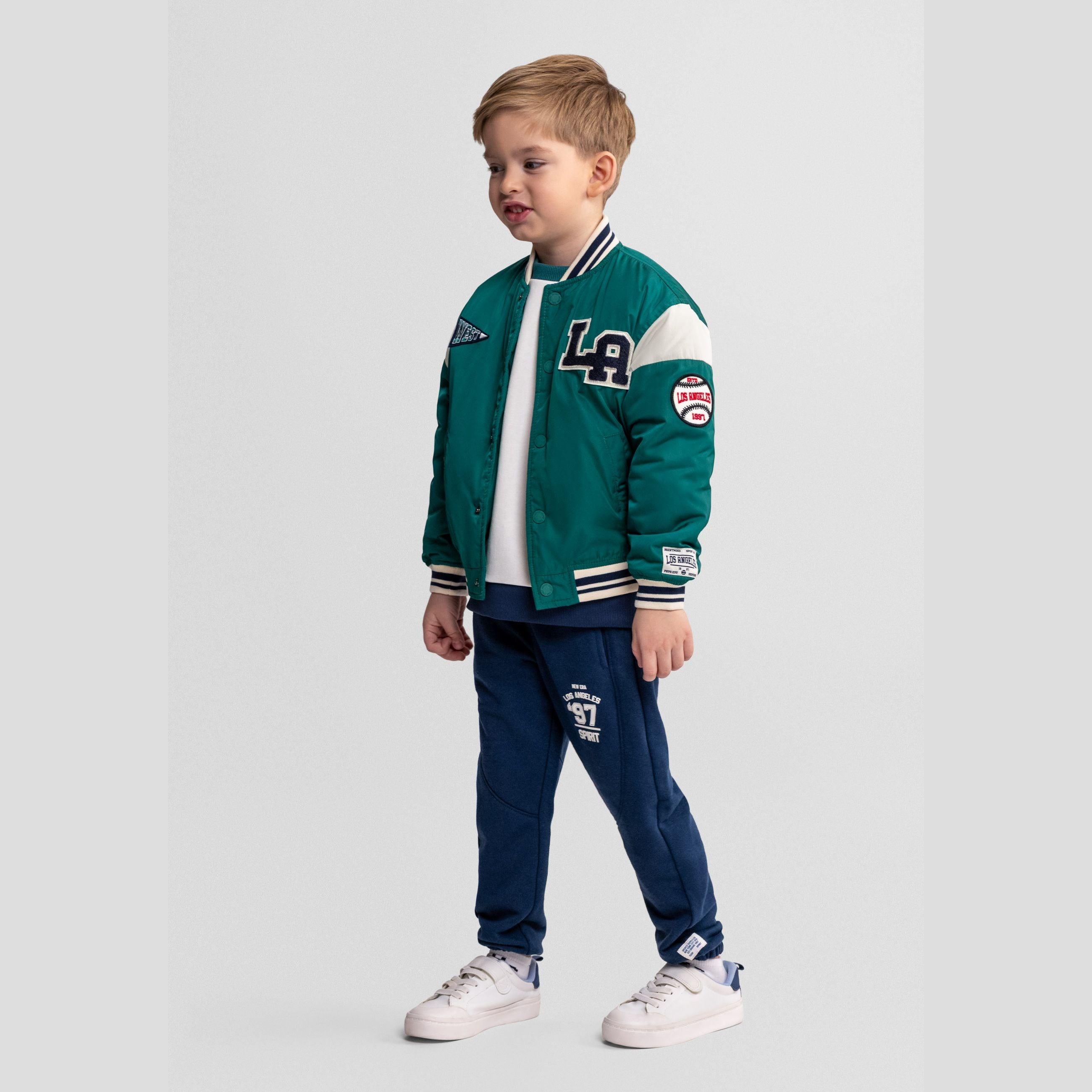 Young boy wearing green varsity bomber jacket with blue joggers and white sneakers, standing against grey background