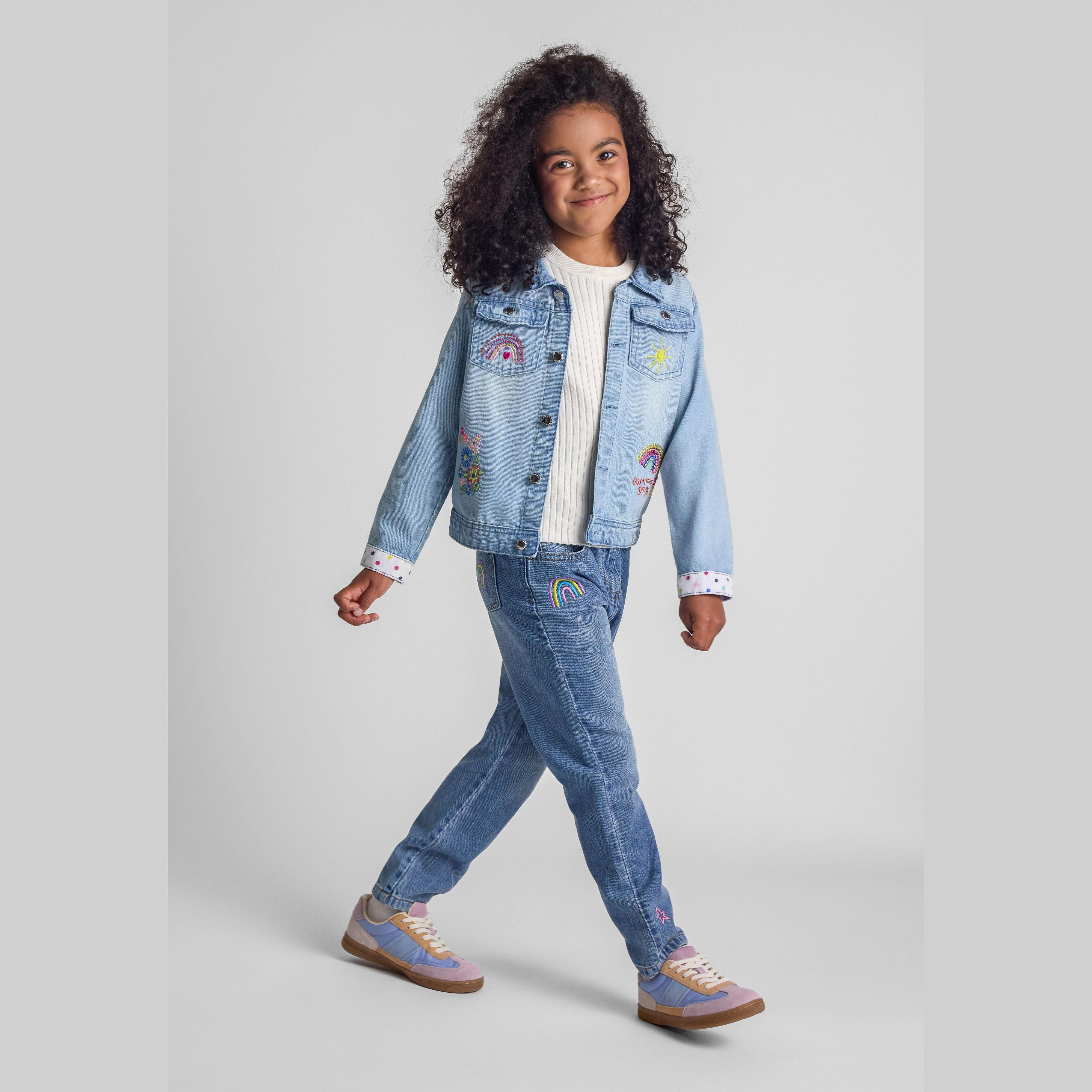 Young girl wearing light blue embroidered denim jacket with jeans, walking pose against grey background