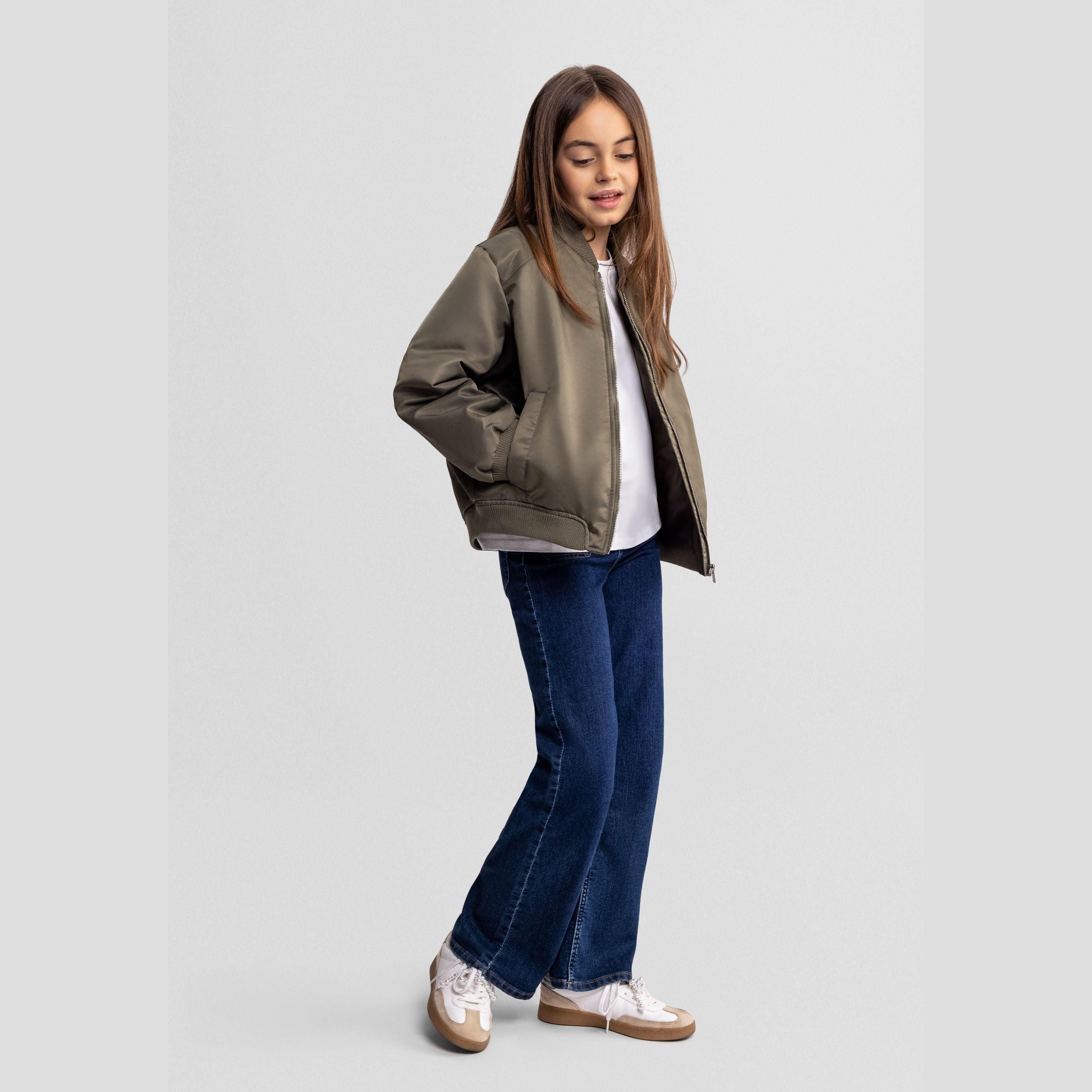 Young girl wearing khaki bomber jacket over white top and dark blue jeans, standing against light grey background