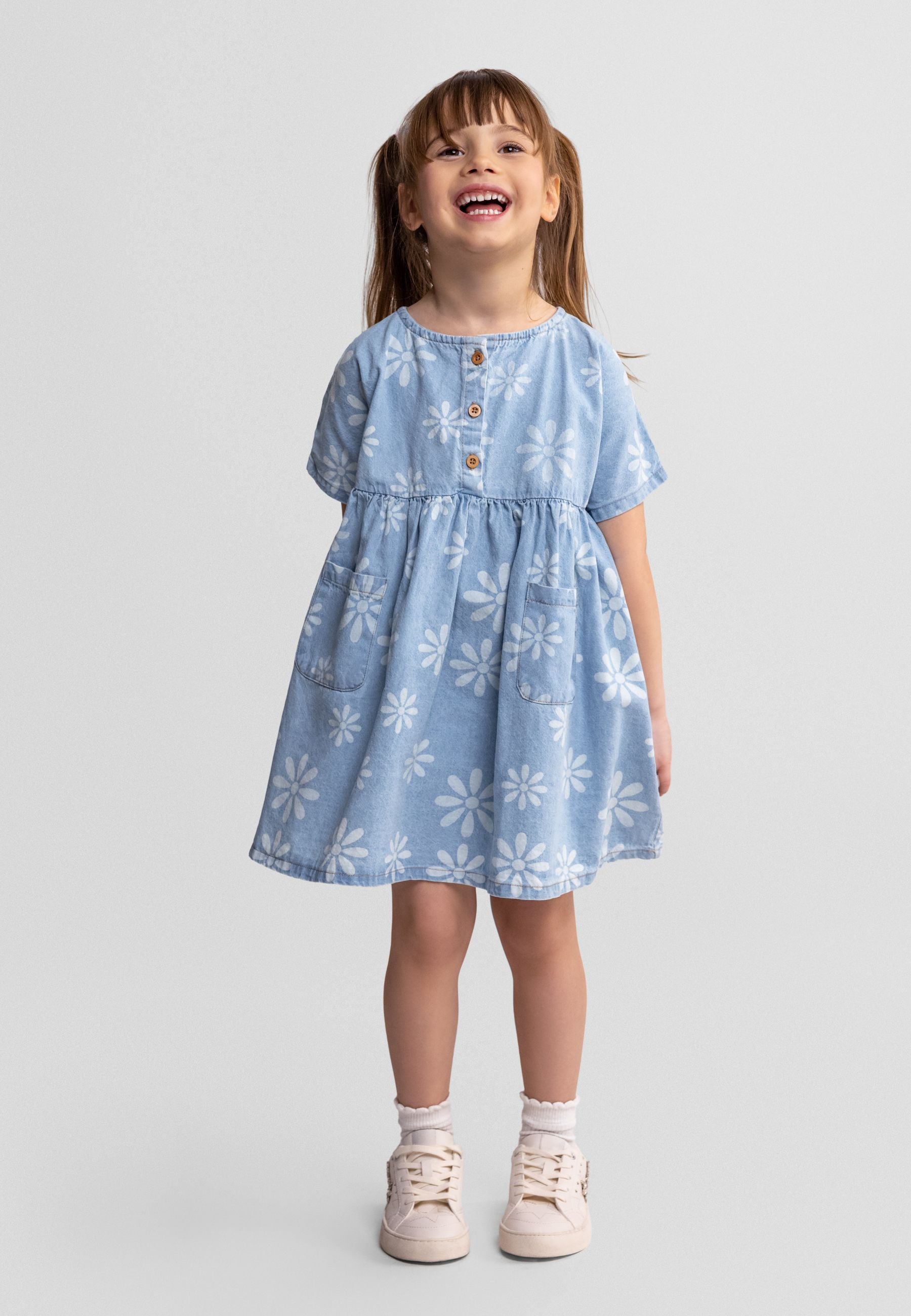 Smiling young girl wearing light blue chambray dress with white daisy print, short sleeves, wooden button front, standing against white background