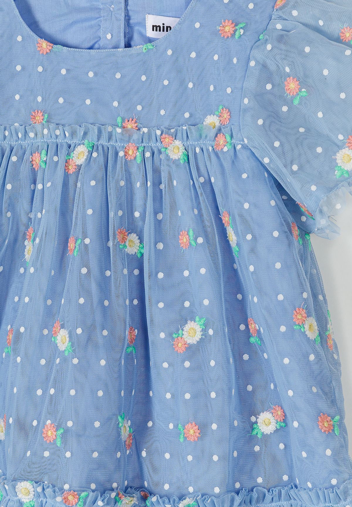 Close-up detail of blue tulle fabric showing white polka dots, coral and green floral embroidery with Minoti label visible