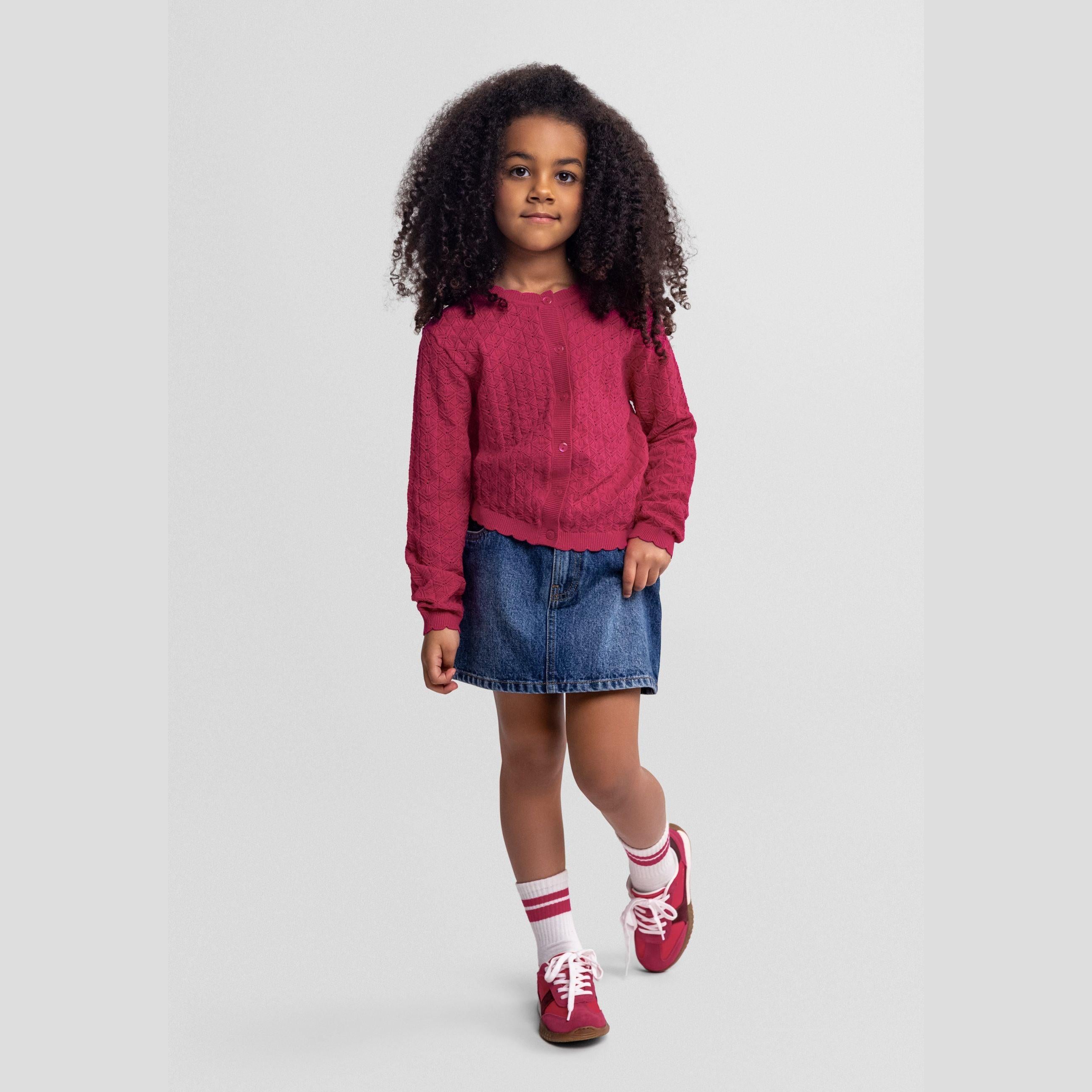Young girl wearing pink diamond knit cardigan over denim skirt, full body front view against grey background