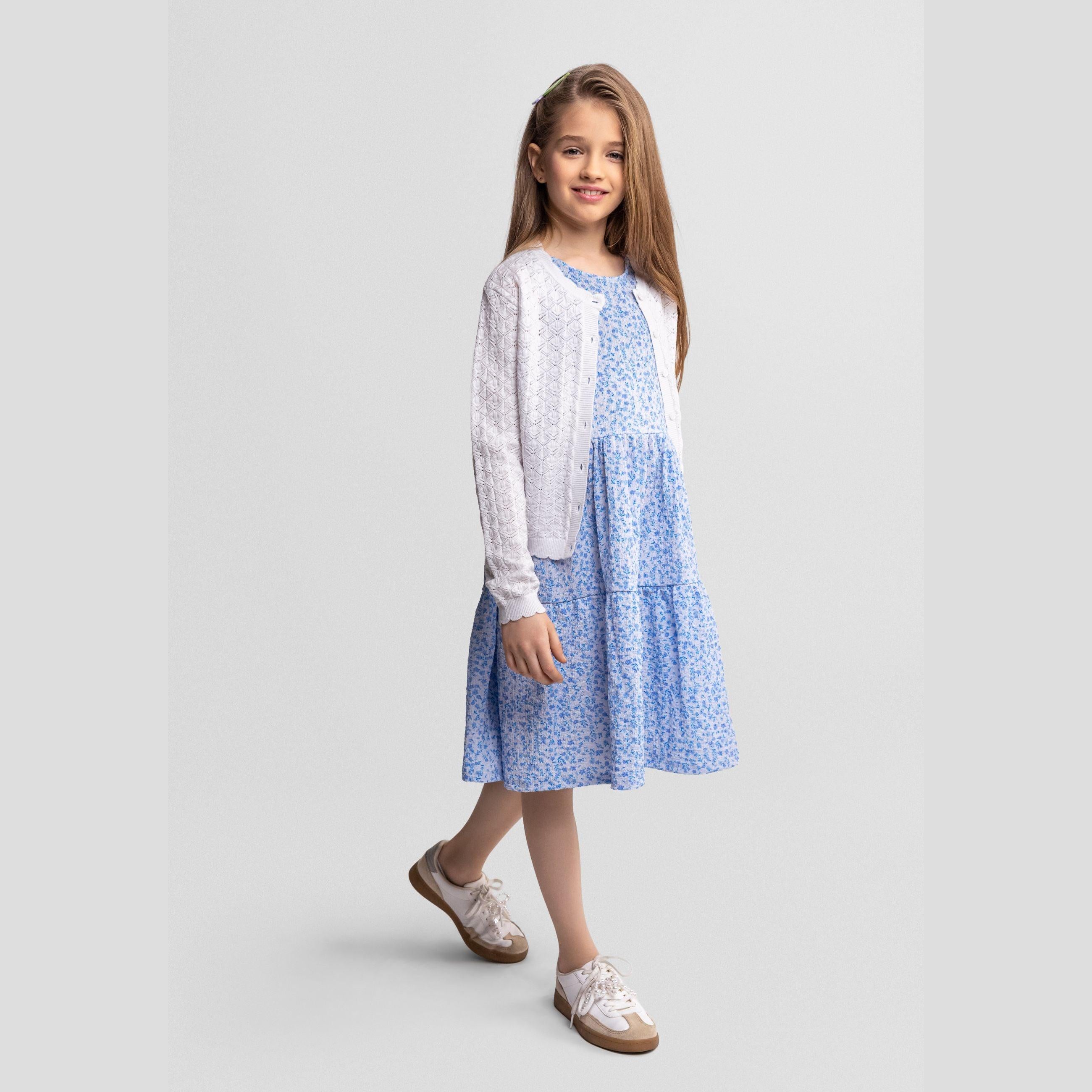 Young girl wearing white wavy trim cardigan over blue floral dress, standing against light grey background