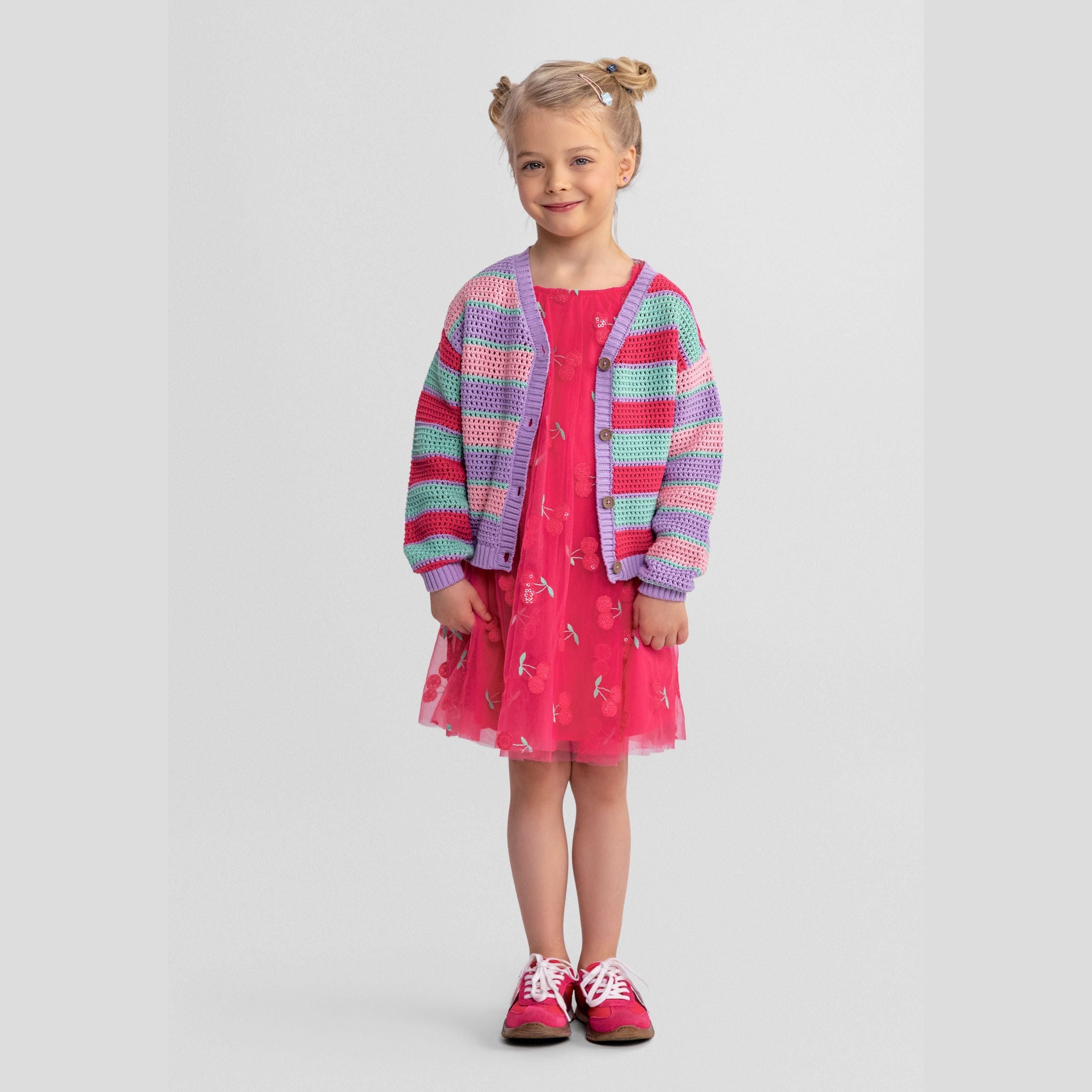 Young blonde girl wearing striped cardigan in lilac, pink and mint over pink tulle dress, smiling at camera against light grey background