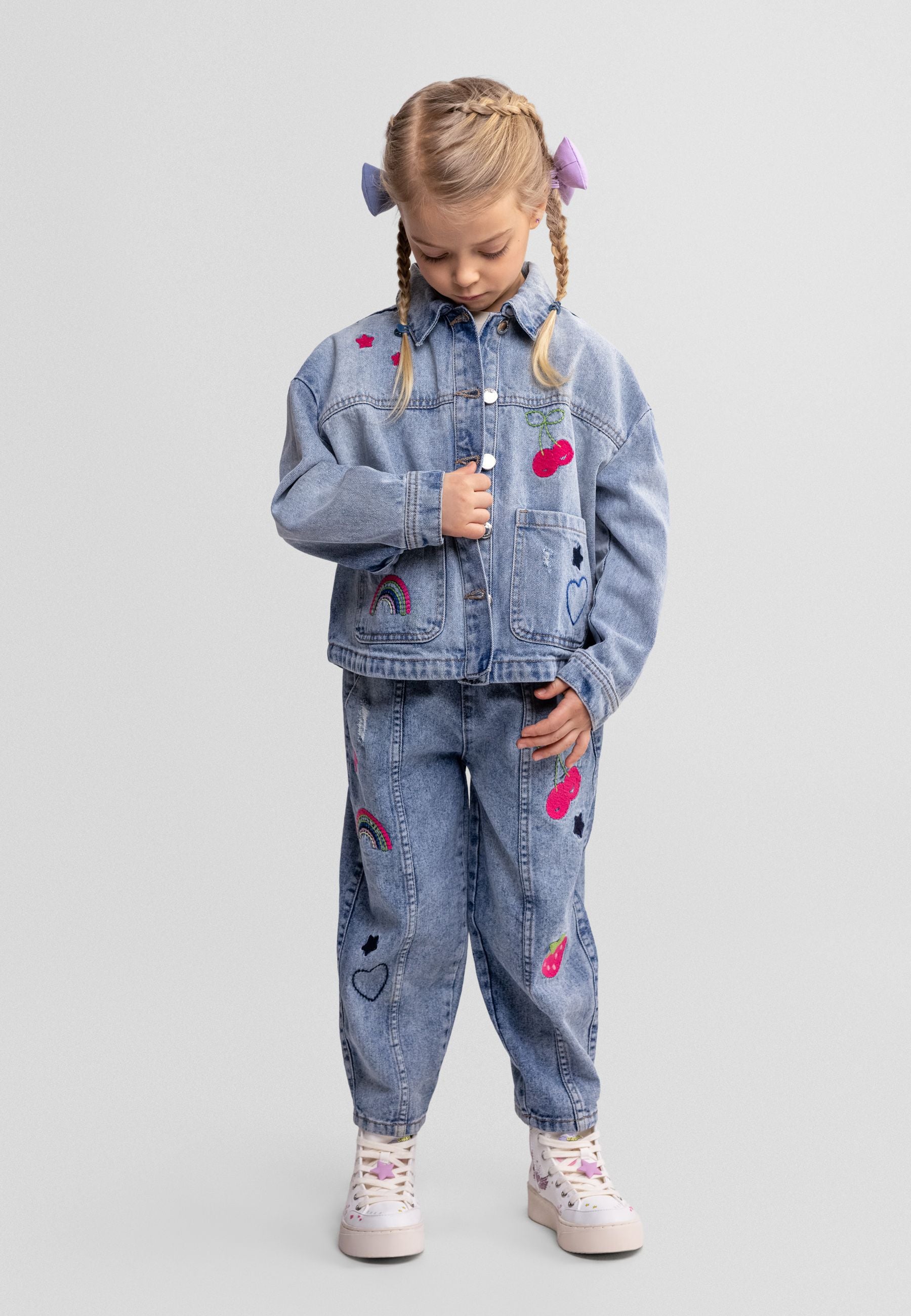 Young girl wearing light blue denim jacket and matching jeans with embroidered cherry and rainbow motifs, styled with braided hair and colourful hair accessories against neutral background