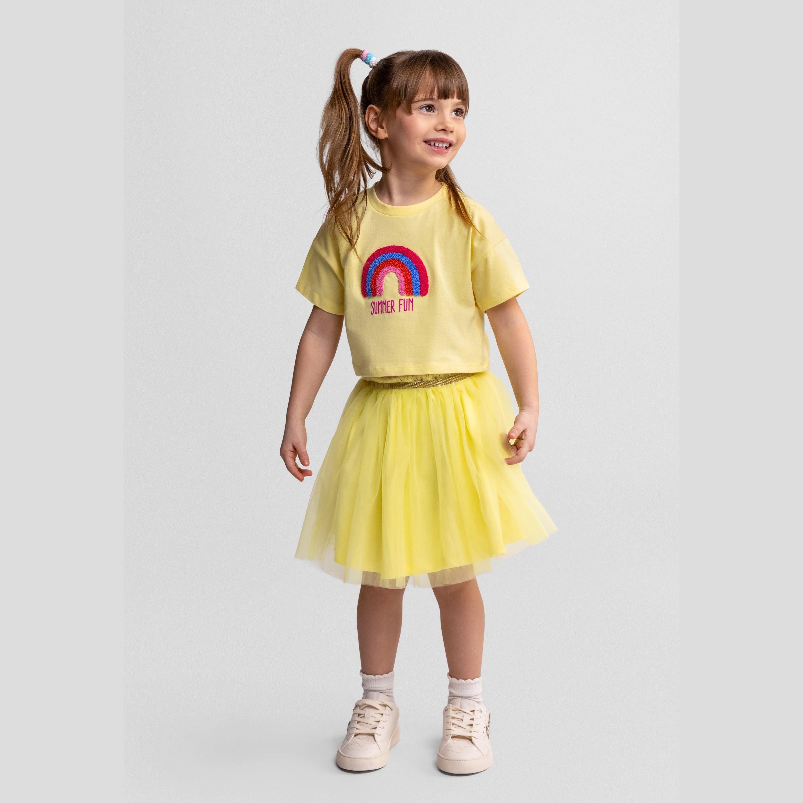 Young girl wearing light yellow tulle skirt with rainbow t-shirt, standing against white background, full body pose showing the skirt's flared silhouette