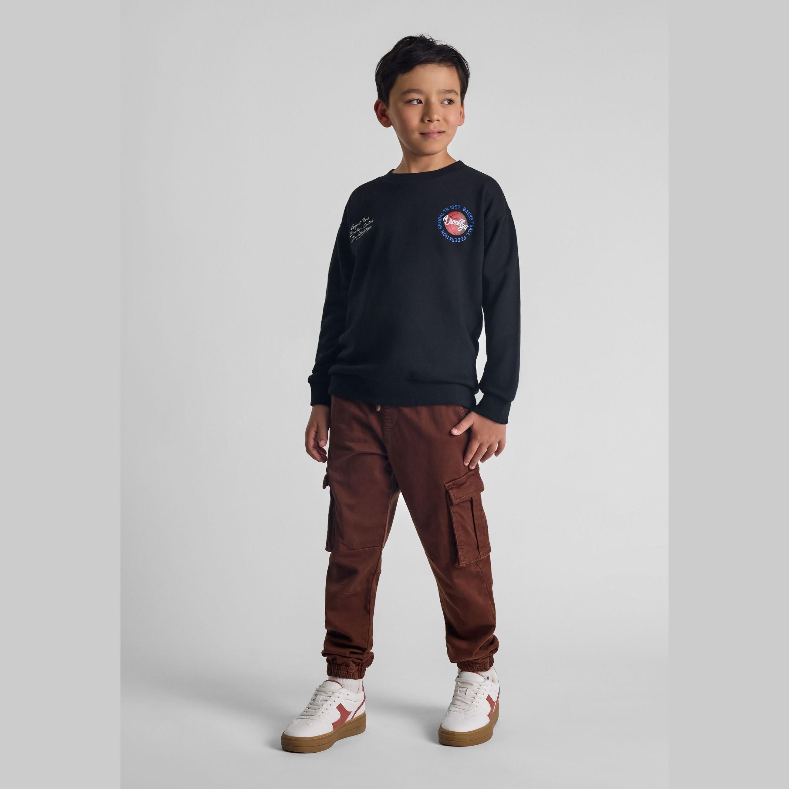 Young boy wearing navy sweatshirt and brown cargo trousers with side pockets, standing against grey background