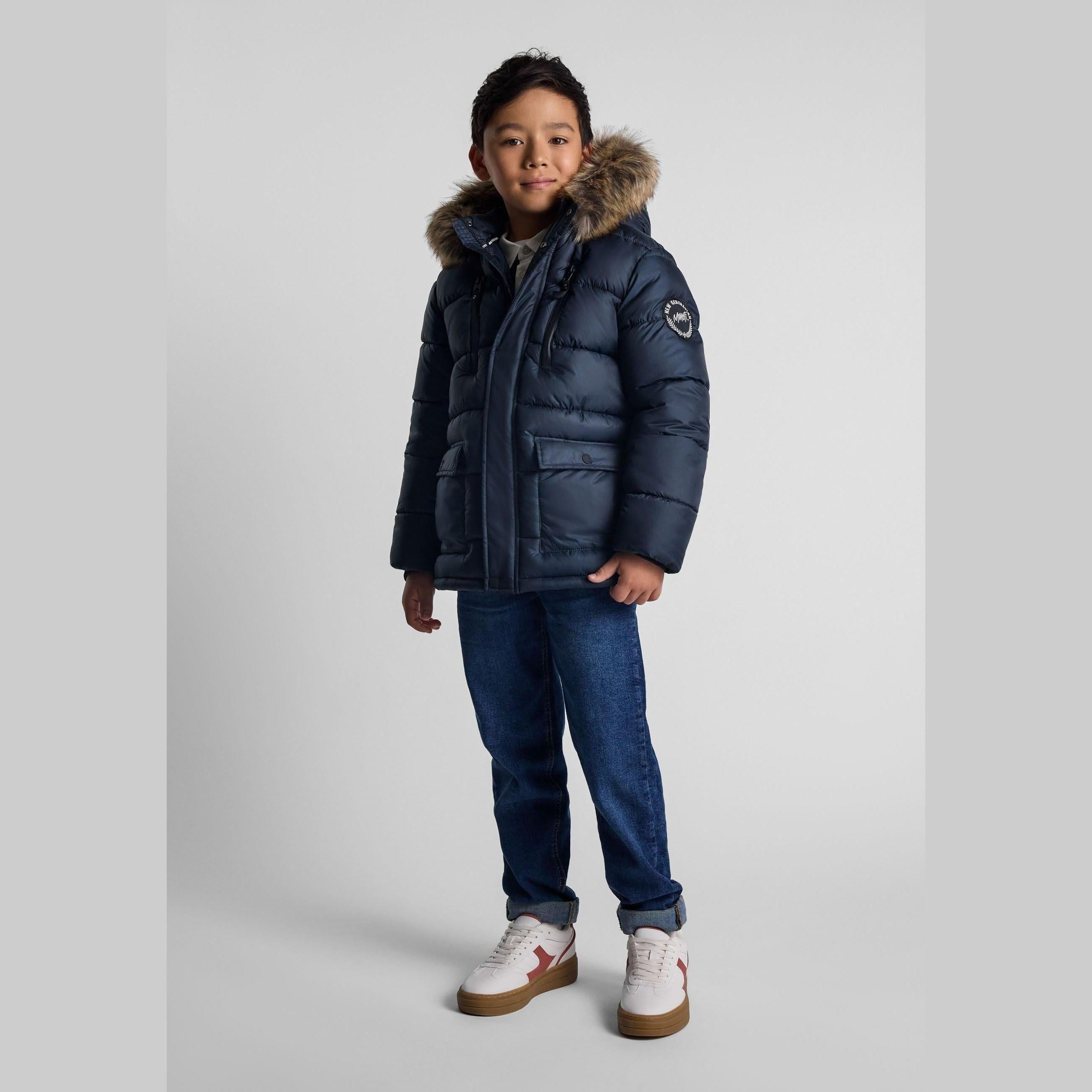 Young boy wearing navy quilted jacket with faux fur hood, paired with jeans and white trainers, standing against light grey background