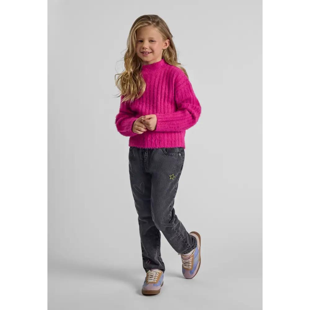 Young girl wearing bright pink fluffy high neck jumper with dark jeans, standing against white background in casual pose