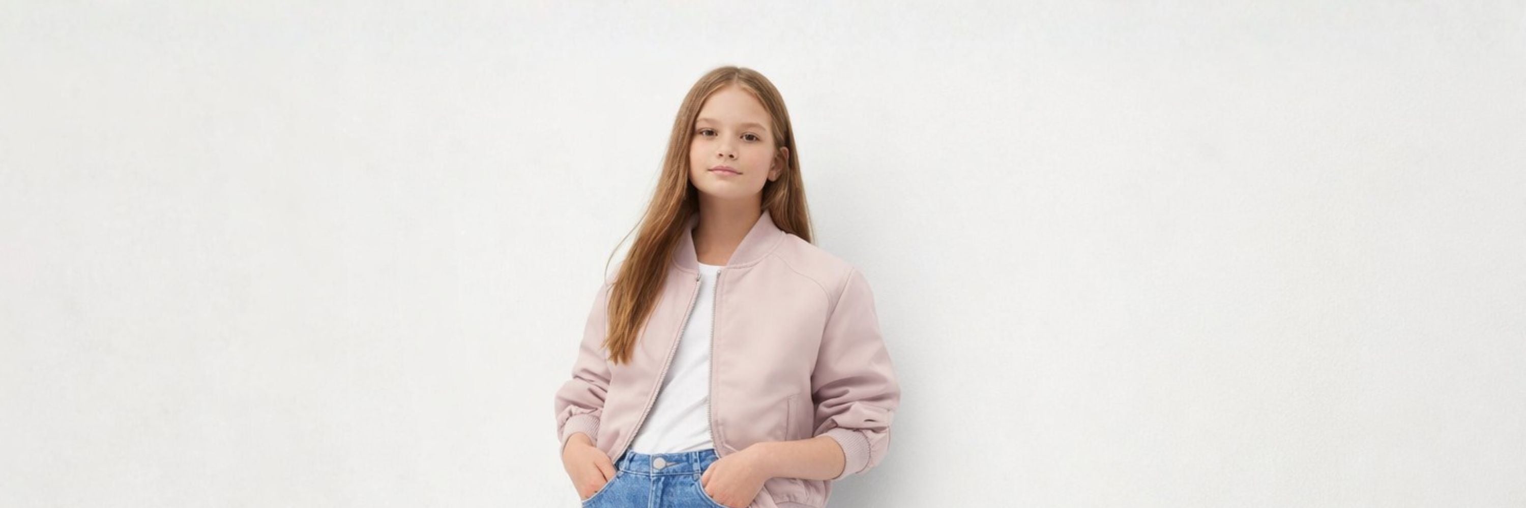A girl in a light pink jacket stands against a clean, minimal background, posing with a calm and confident expression.