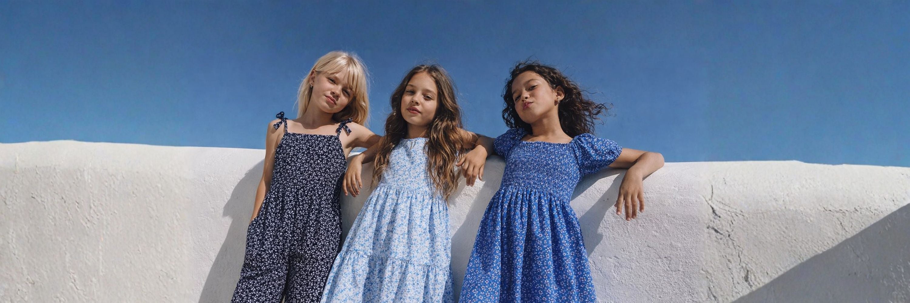 Three girls in lightweight summer dresses lean casually against a white wall, posing confidently under a clear blue sky.