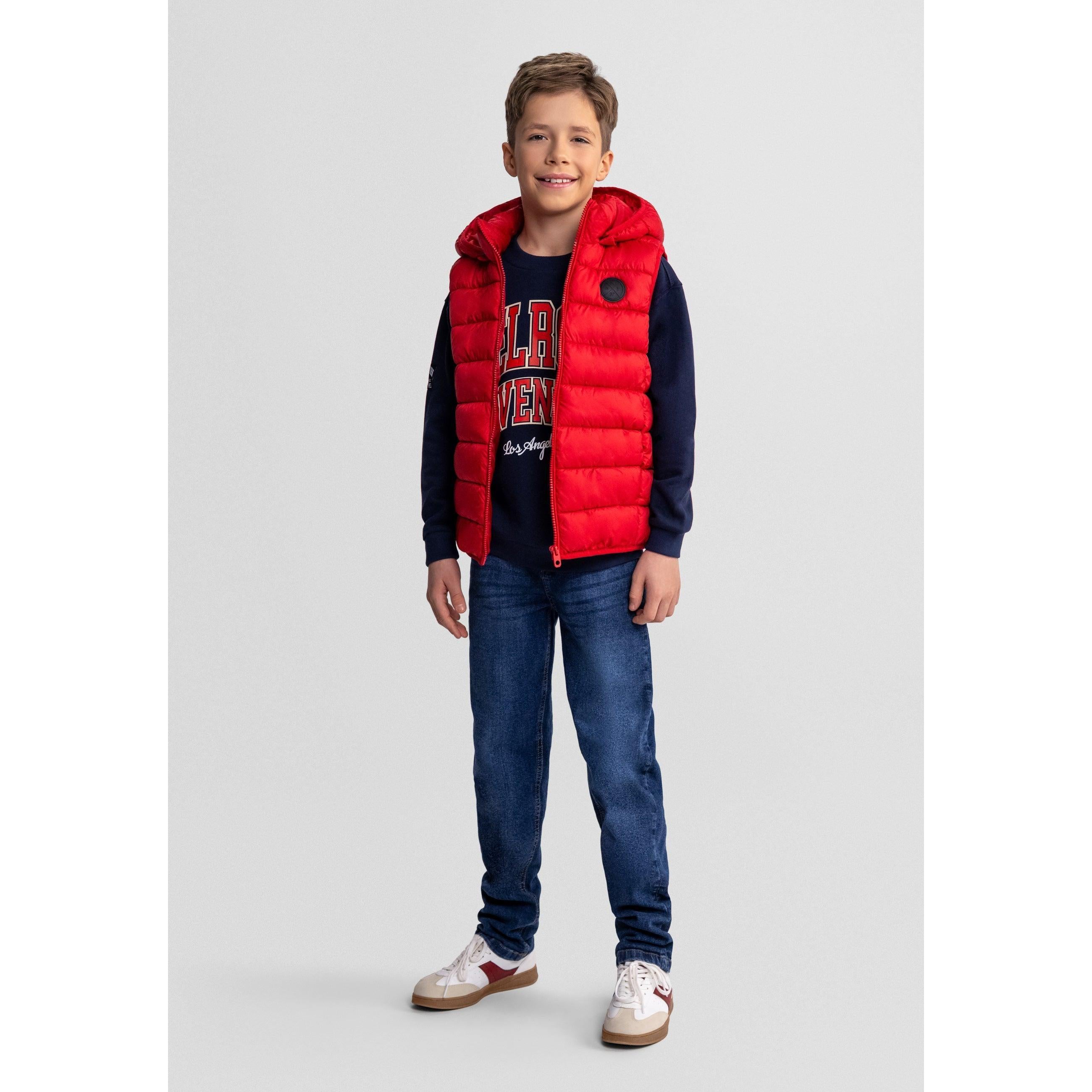 Boy wearing red padded gilet with detachable hood over navy sweatshirt and jeans, standing against grey background