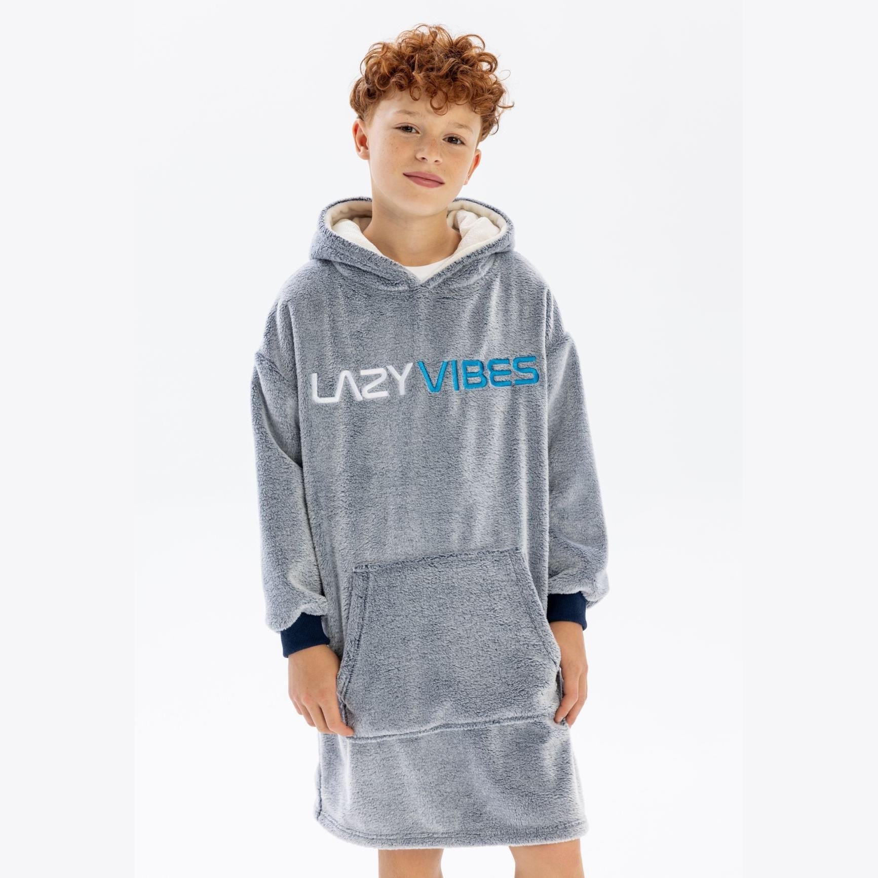 Boy with curly hair wearing grey oversized fleece hoodie with LAZY VIBES text, standing against white background with hands in kangaroo pocket