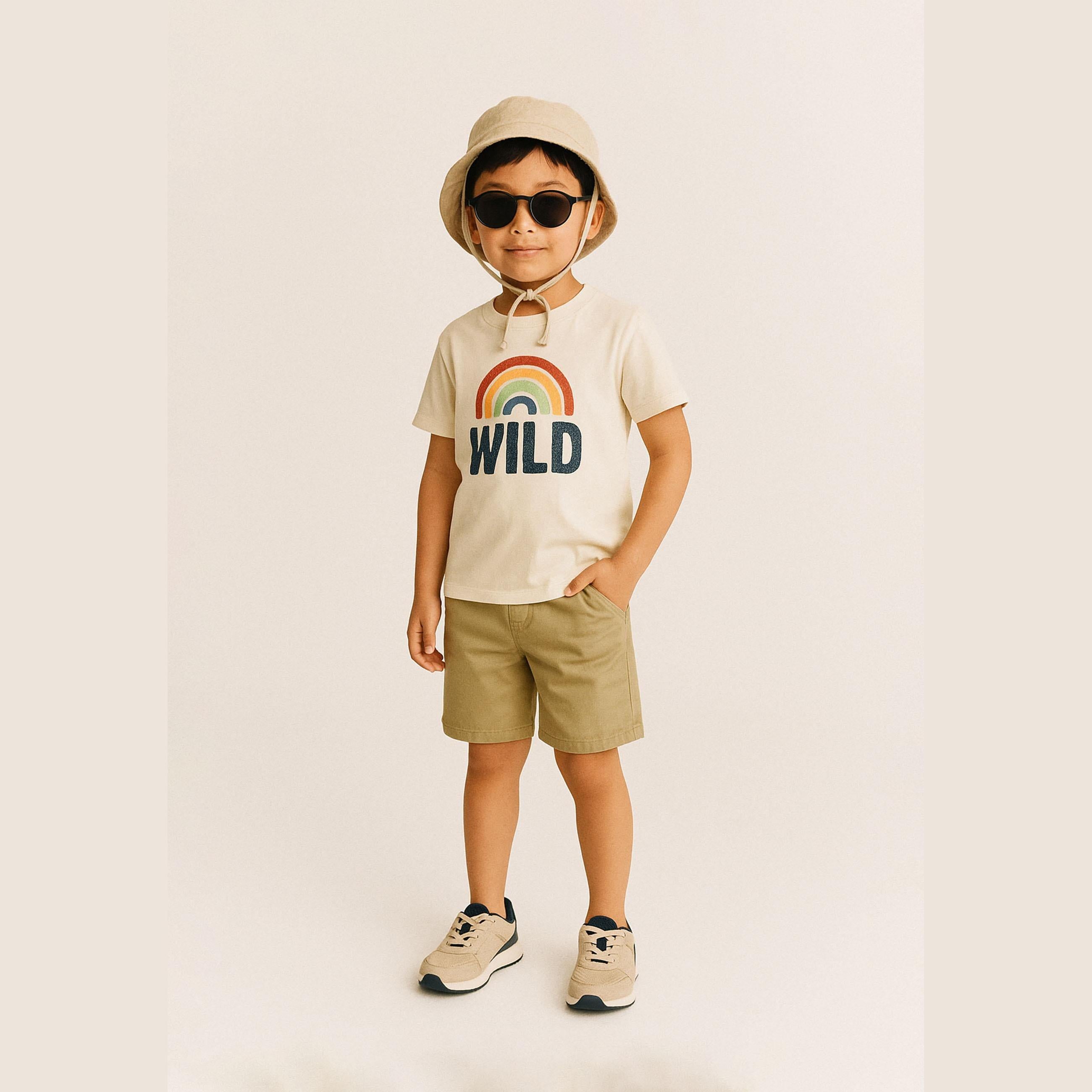 Boy wearing beige chino shorts with cream WILD rainbow t-shirt, sunhat and sunglasses against beige background