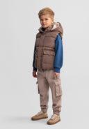 Young boy wearing a beige hooded gilet with teddy fleece lining and patch pockets, paired with casual beige pants and sneakers.