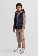Smiling boy wearing a black quilted sleeveless vest with hood over beige cargo pants and sneakers, casual kids fashion design.