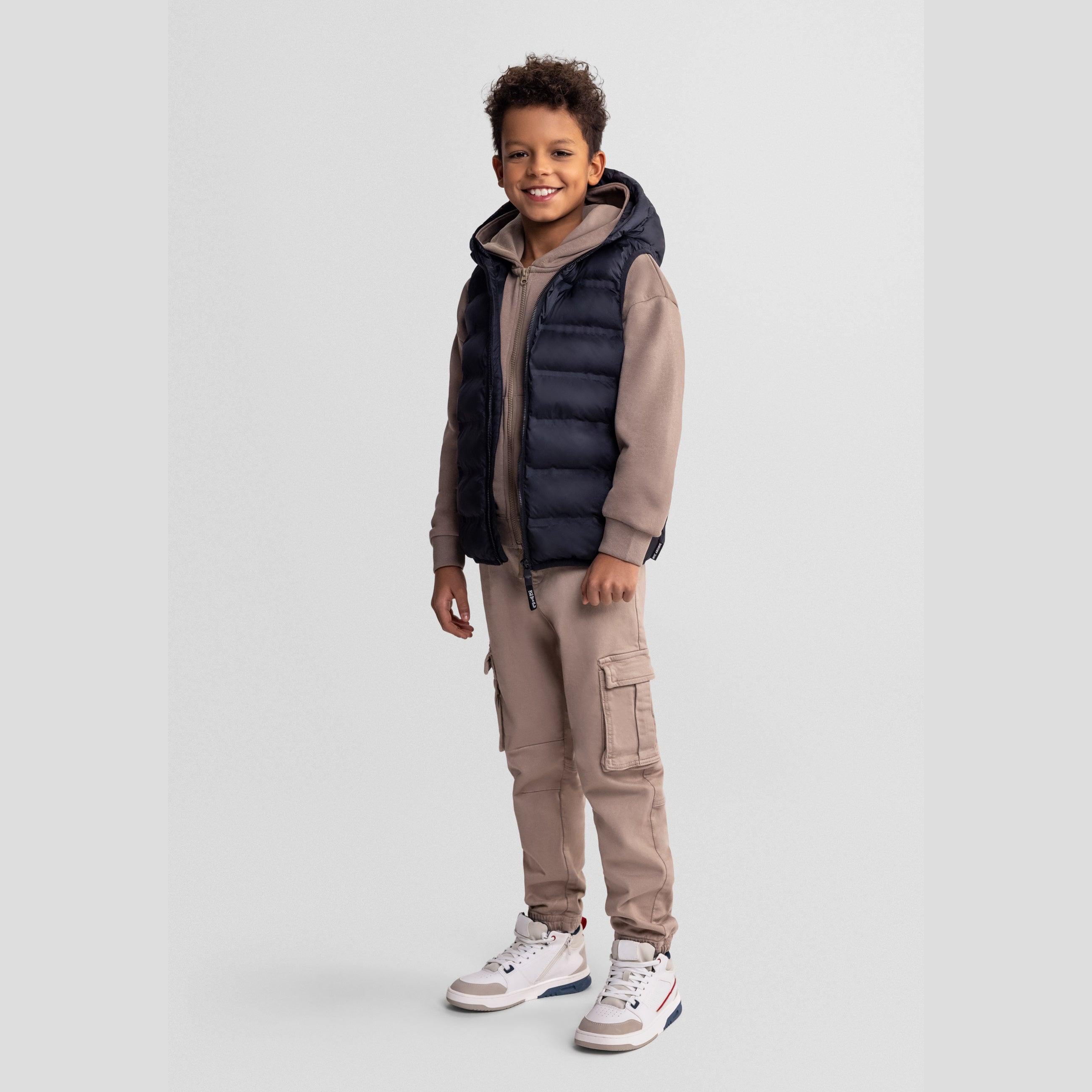 Boy wearing black quilted hooded waistcoat over beige hoodie and cargo trousers, standing against light grey background